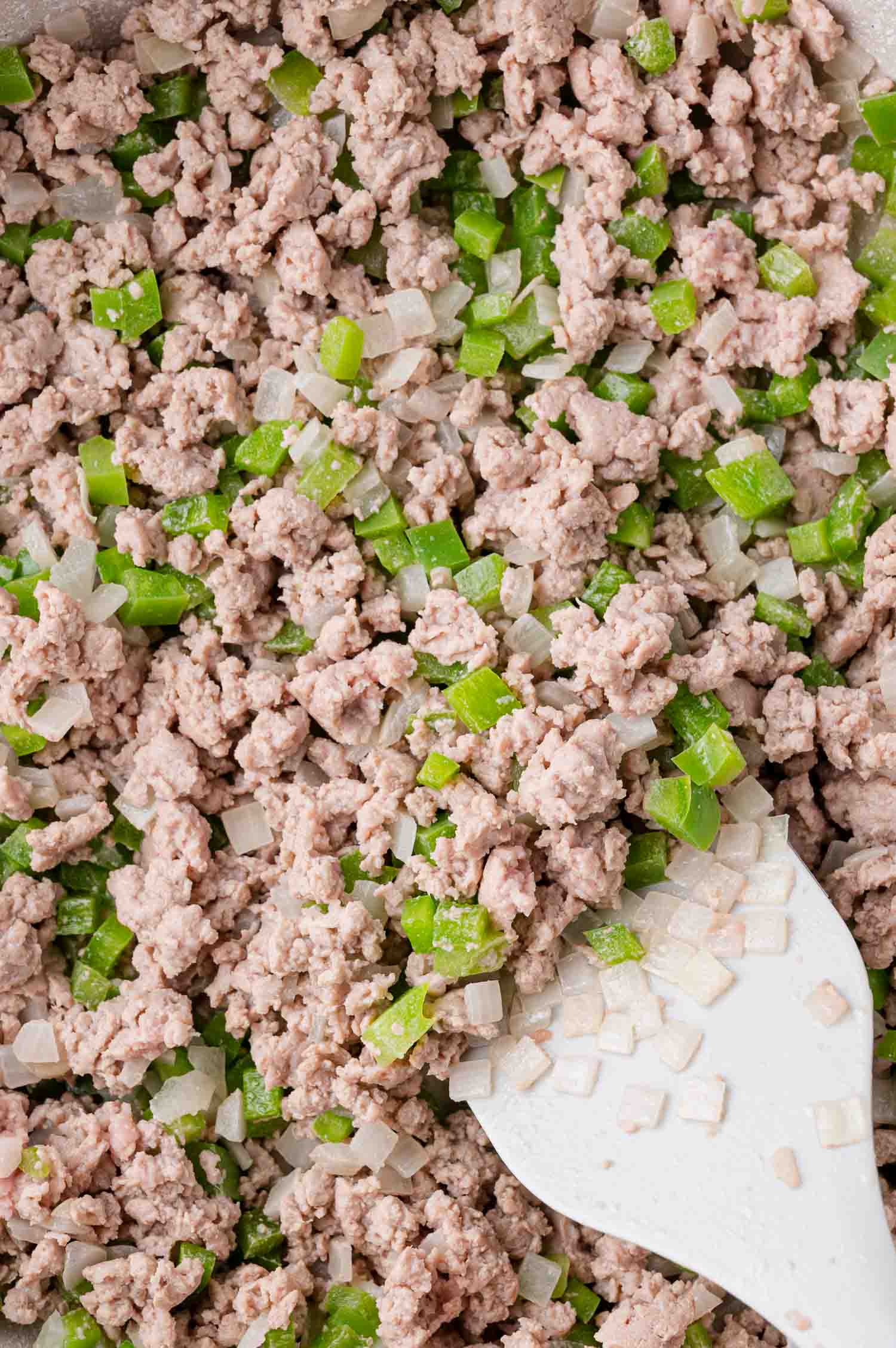 Cooked ground meat mixed with diced green bell peppers and onions, being stirred with a white spatula in a pan.