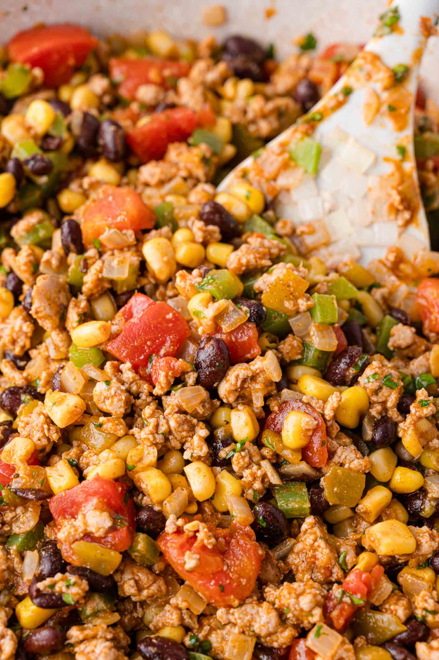 A close-up of a mixture containing ground meat, black beans, corn, diced tomatoes, and chopped green peppers being stirred with a white spatula.