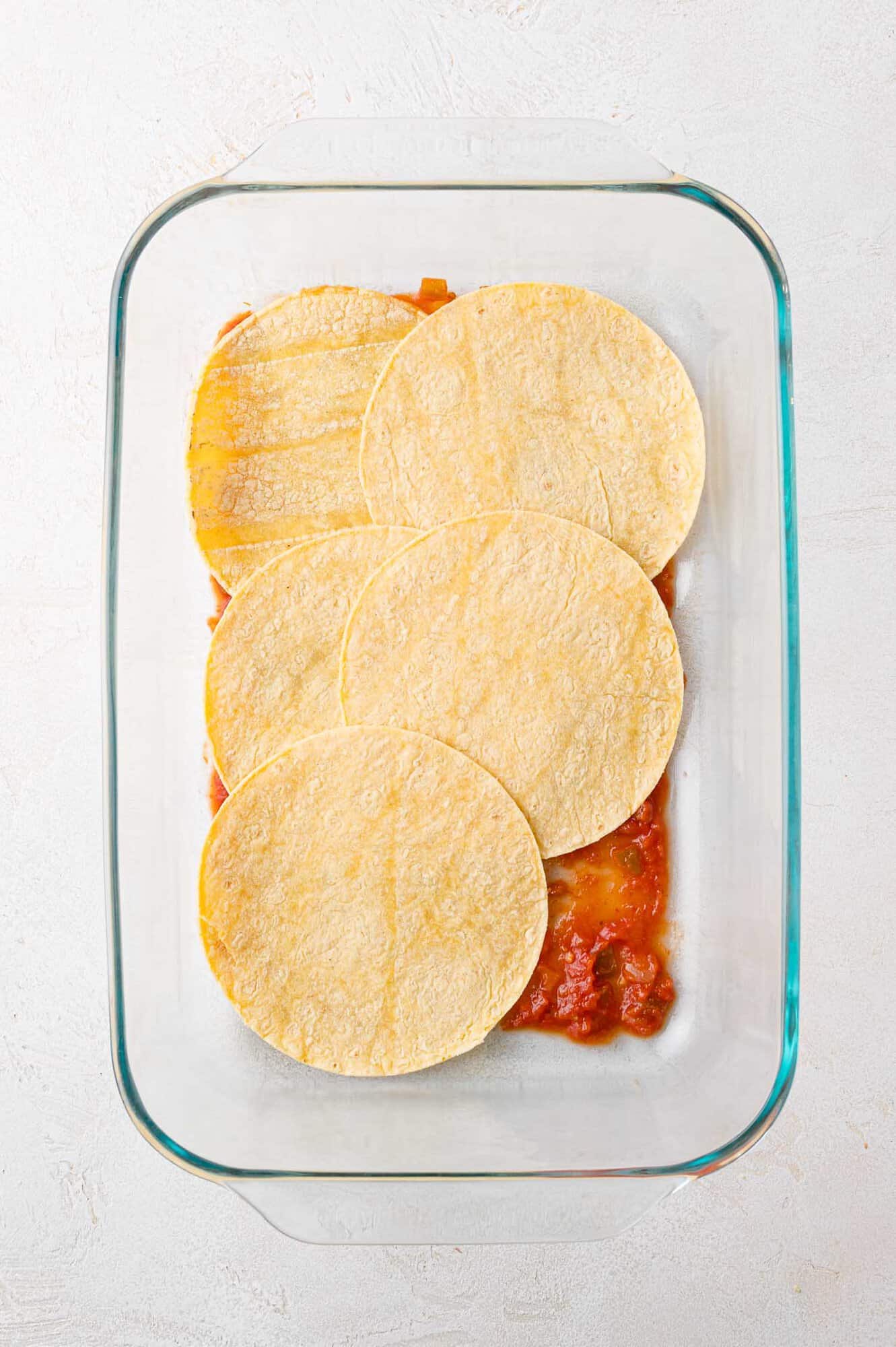 A glass baking dish with a layer of salsa and whole corn tortillas neatly arranged on top.