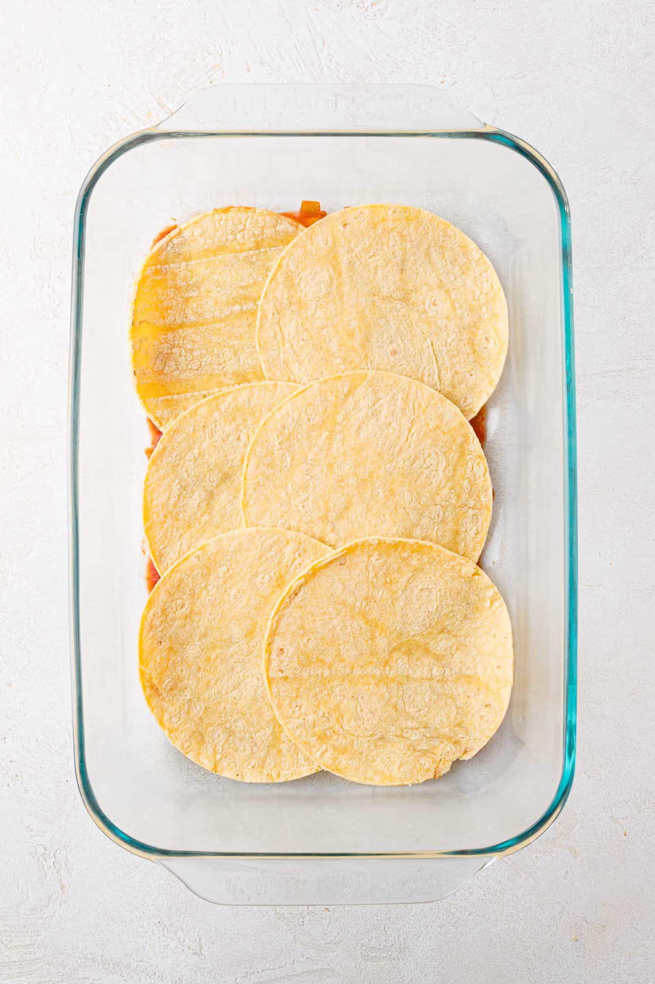 A glass baking dish with six overlapping corn tortillas laid flat, covering a layer of ingredients underneath.