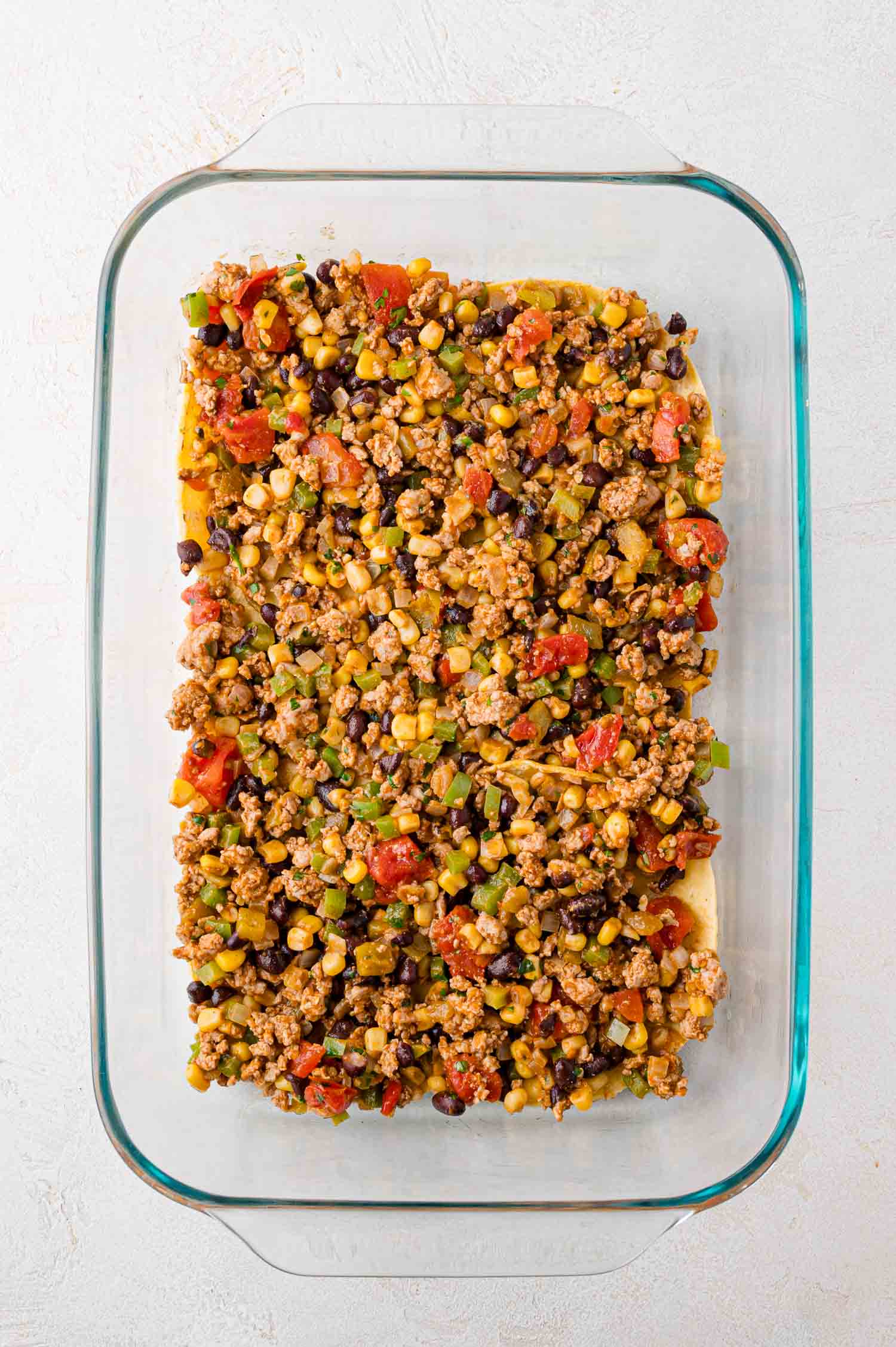 A glass baking dish filled with a mixture of ground meat, black beans, corn, diced tomatoes, and chopped vegetables on a white surface.