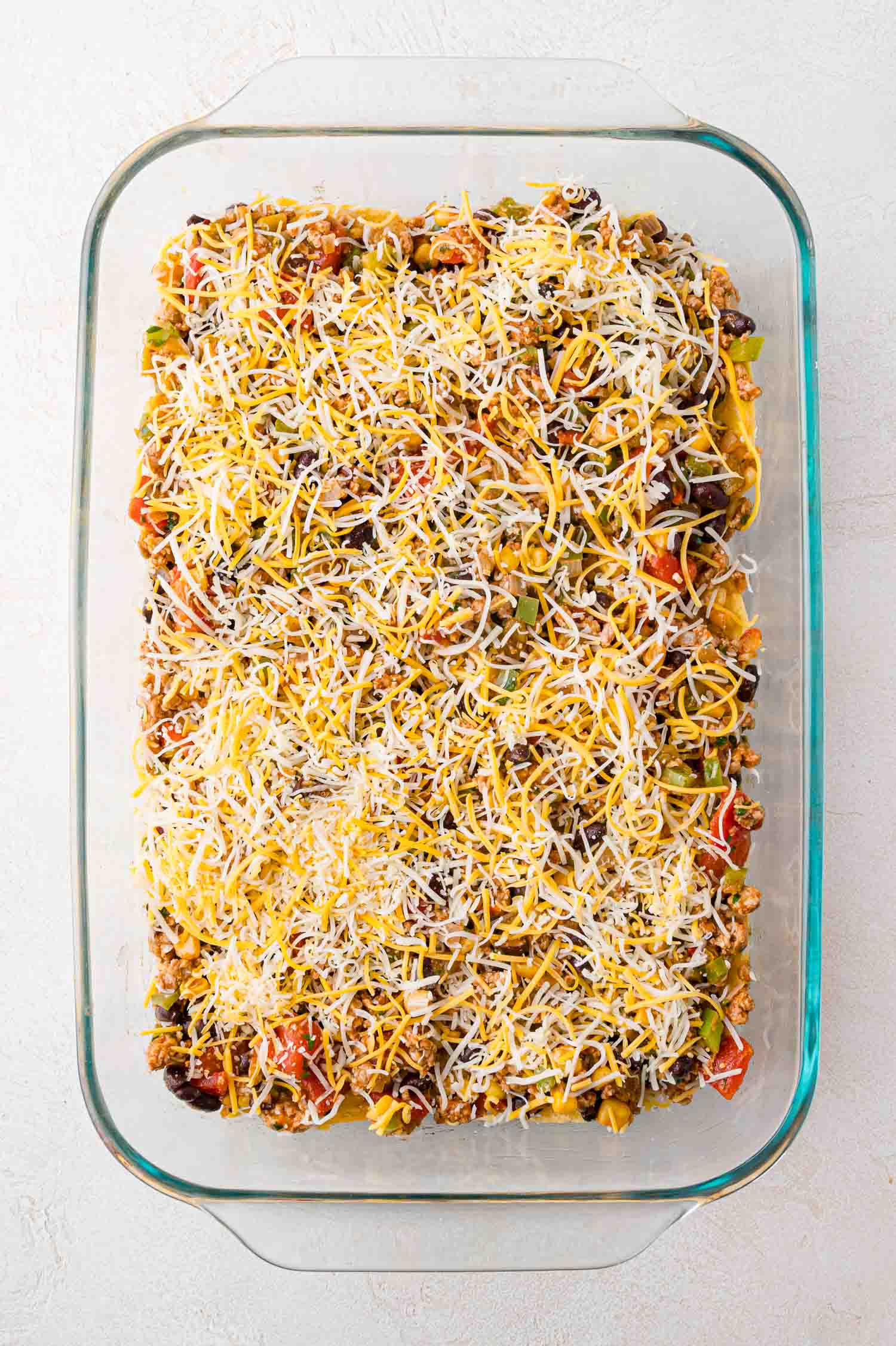 A glass baking dish filled with an unbaked casserole containing ground meat, vegetables, and topped with shredded cheese.