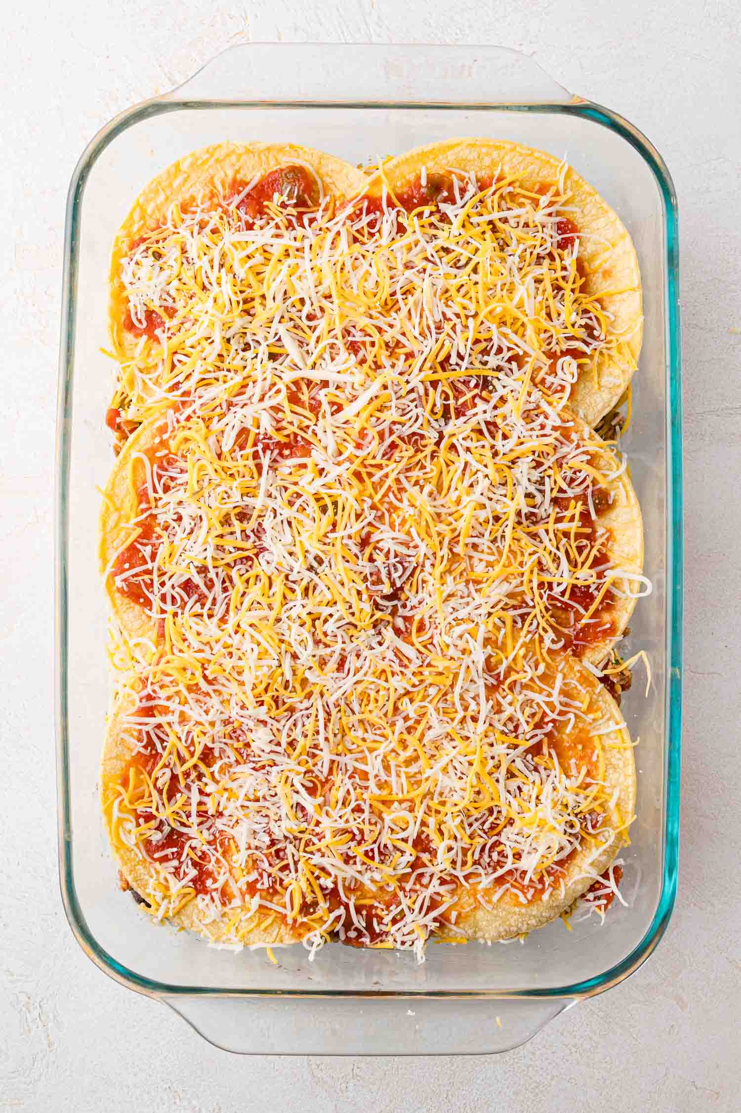 A glass baking dish filled with stacked tortillas, red sauce, and shredded cheese, ready to be baked.