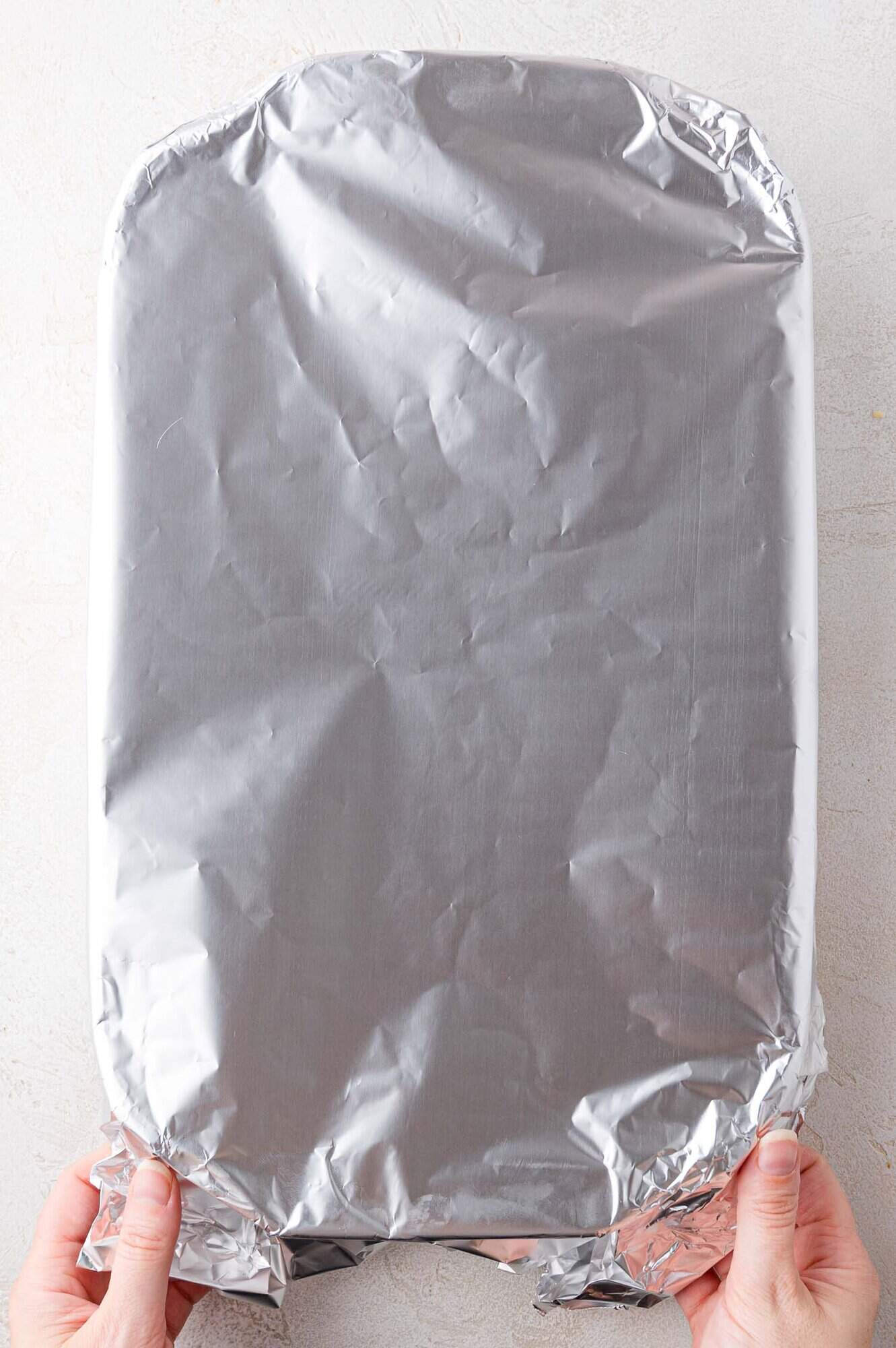 A pair of hands holds a large rectangular baking dish covered with aluminum foil on a light surface.