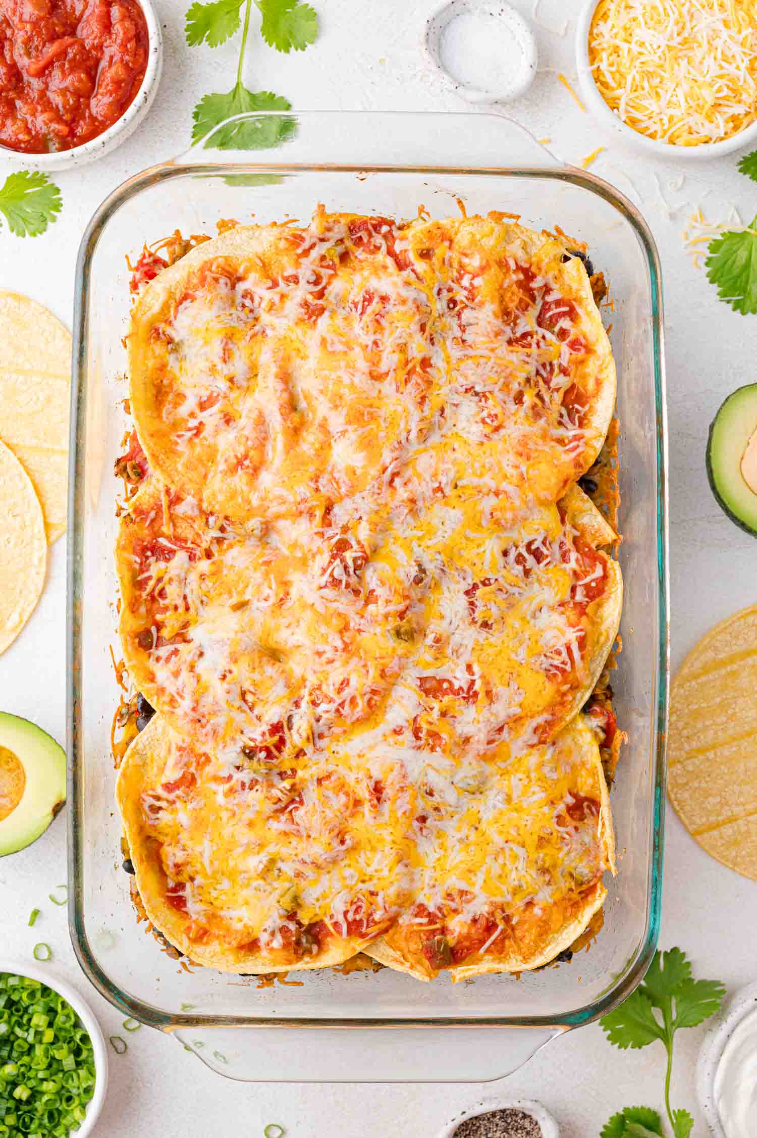 A glass baking dish filled with a layered mexican lasagna topped with melted cheese; surrounded by tortillas, avocado, shredded cheese, and small bowls of salsa and seasonings.