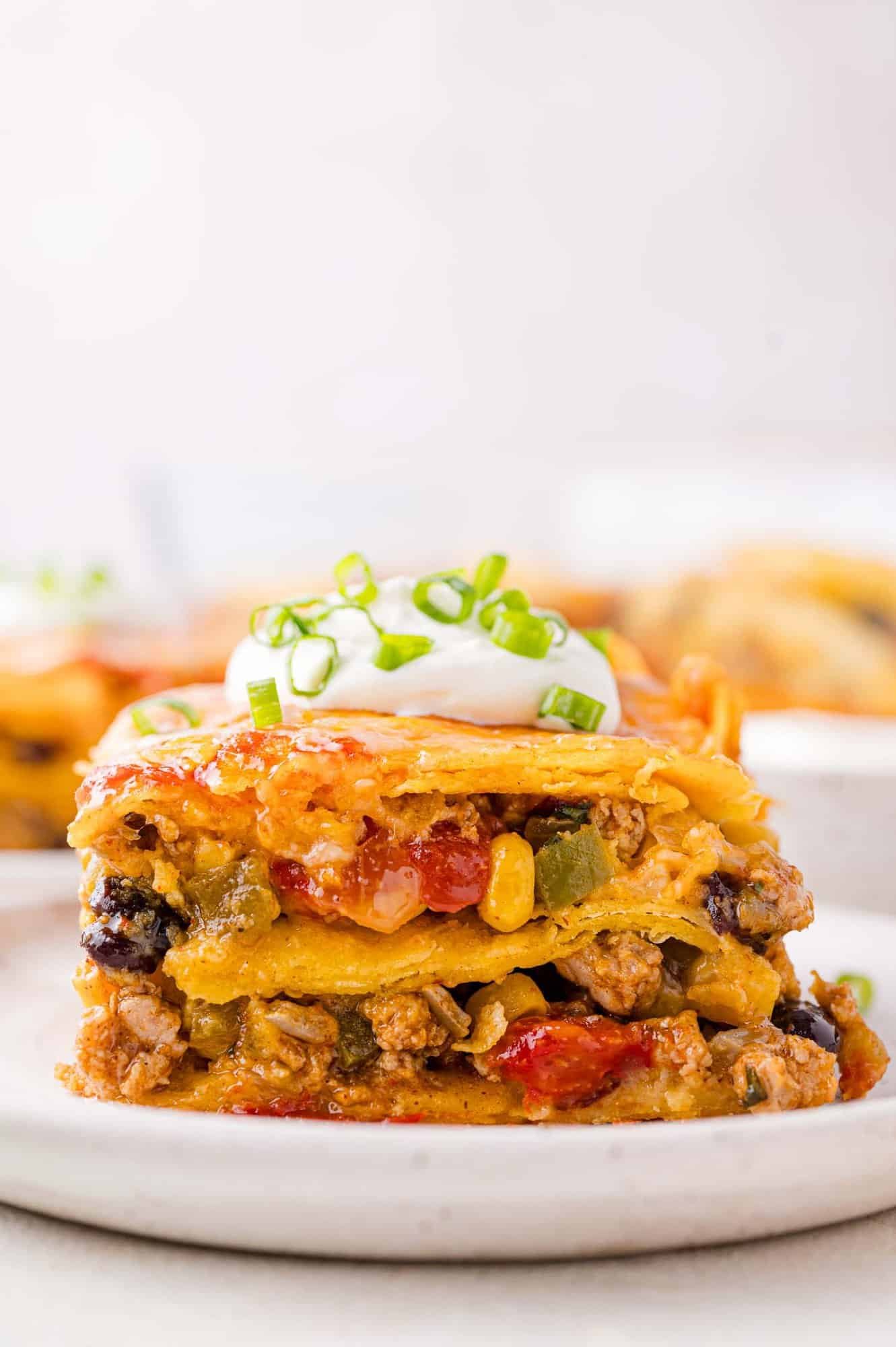 A slice of layered Mexican casserole with ground meat, beans, corn, and vegetables, topped with sour cream and chopped green onions on a white plate.