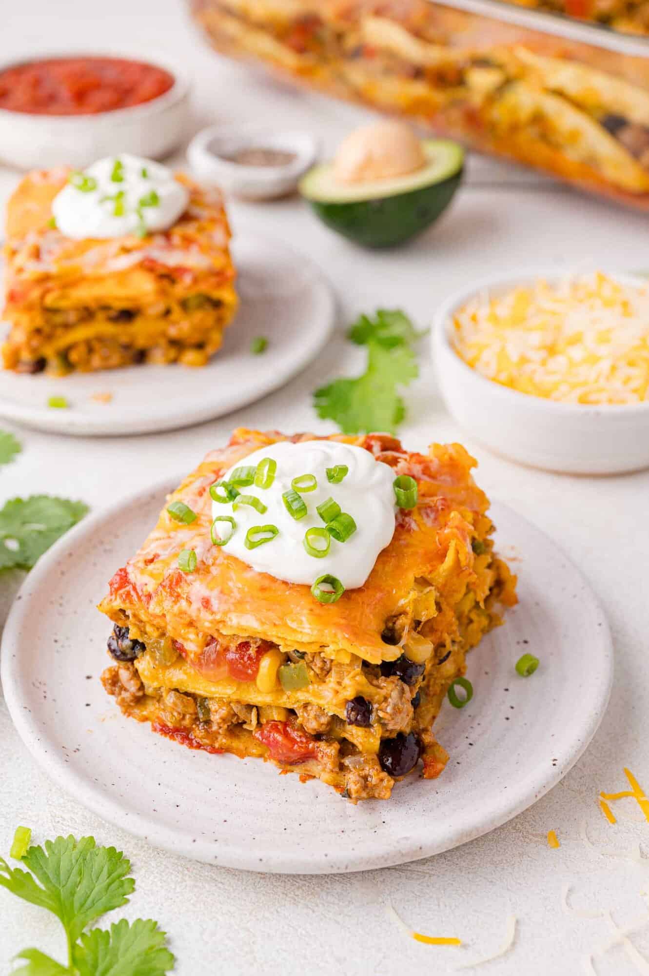 A slice of layered Mexican lasagna topped with sour cream and chopped green onions on a plate, with another serving and ingredients in the background.