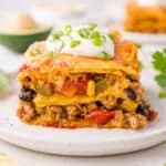 A slice of layered Mexican lasagna with ground meat, beans, cheese, and tomatoes topped with sour cream and chopped green onions on a white plate.