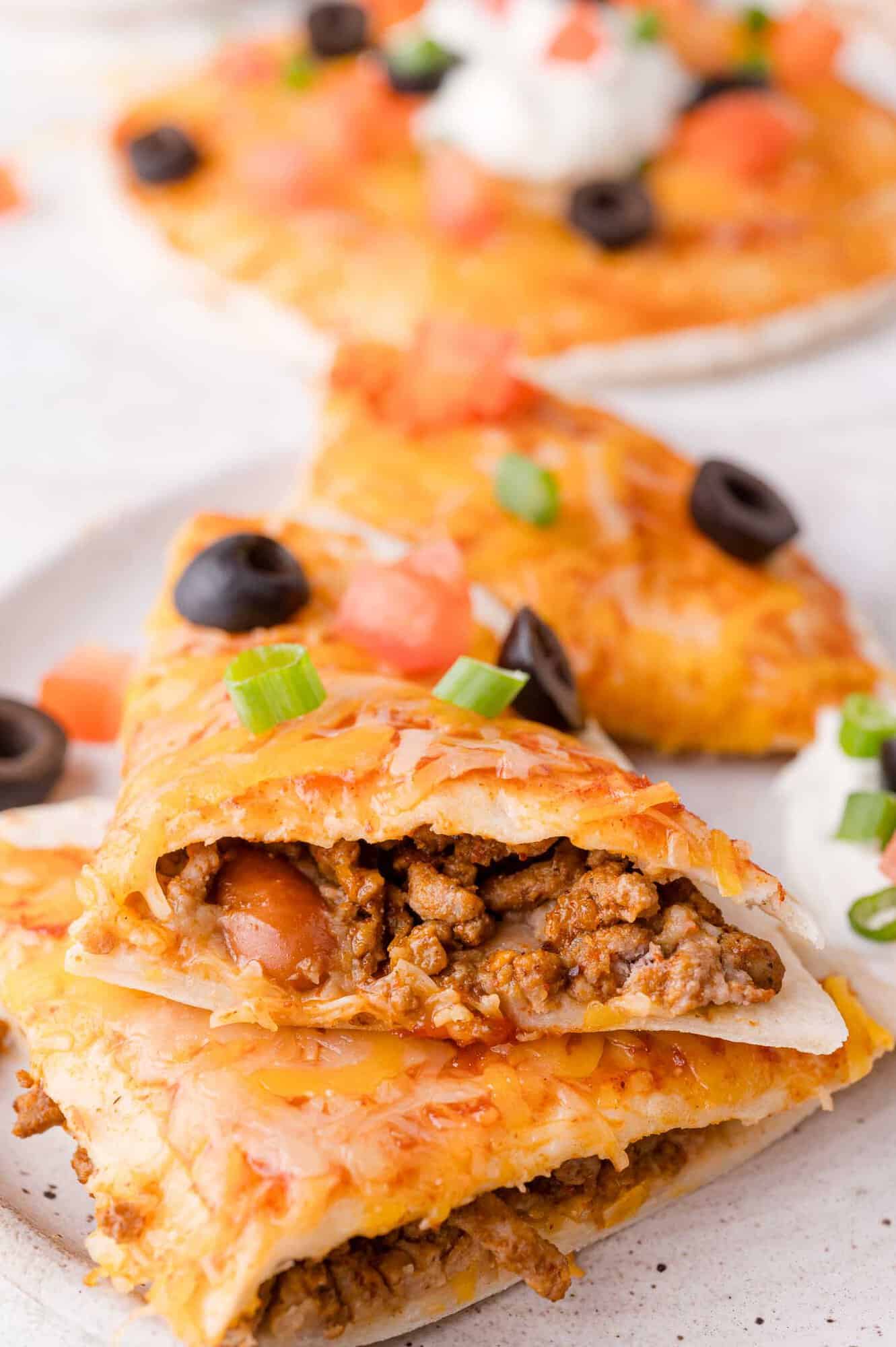 Close-up of a Mexican pizza slice filled with ground beef, topped with melted cheese, olives, tomatoes, sour cream, and green onions. Other slices visible in the background.
