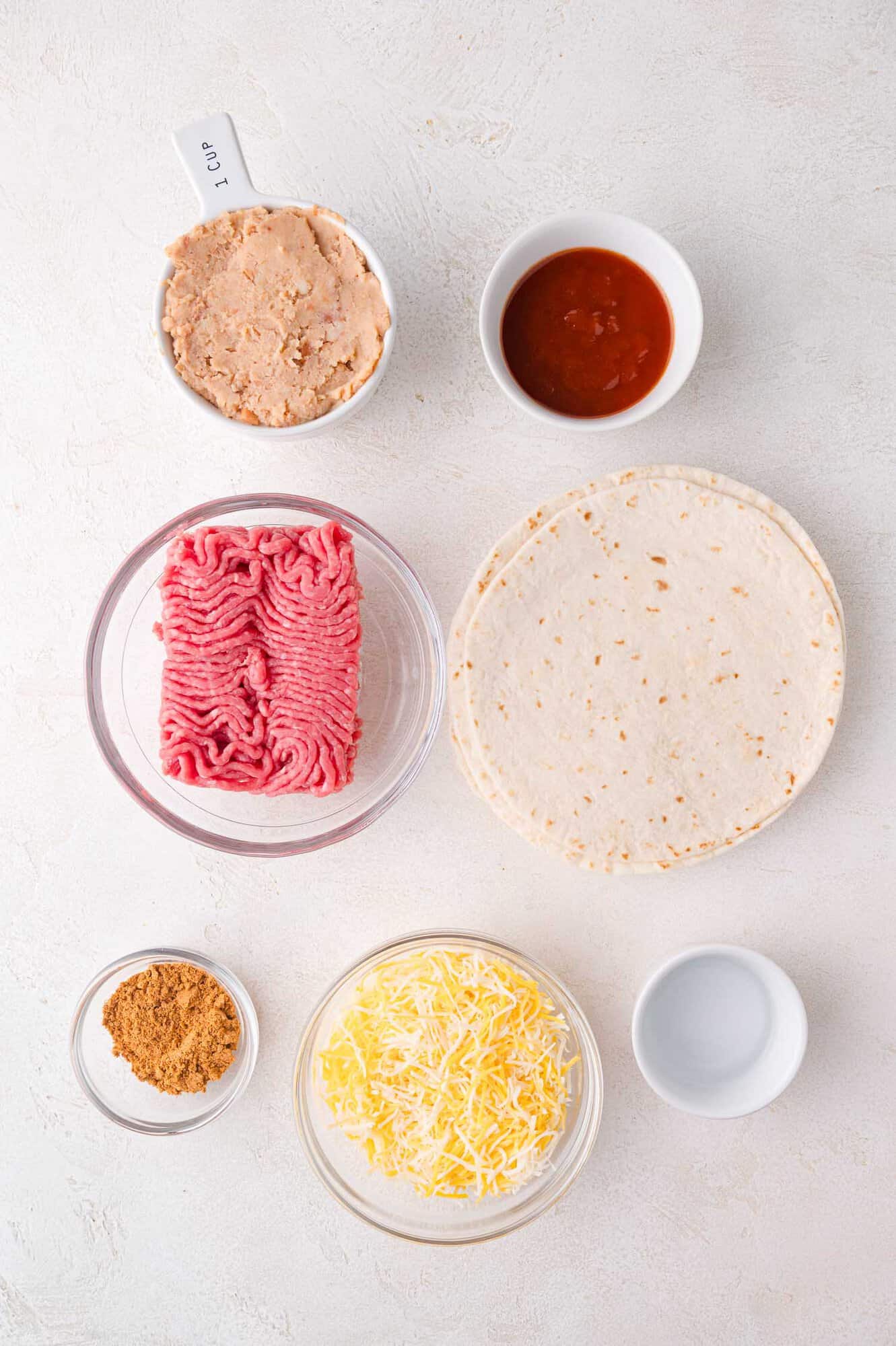 Ingredients arranged on a white surface include raw ground meat, tortillas, shredded cheese, refried beans, salsa, taco seasoning, and an empty measuring cup.