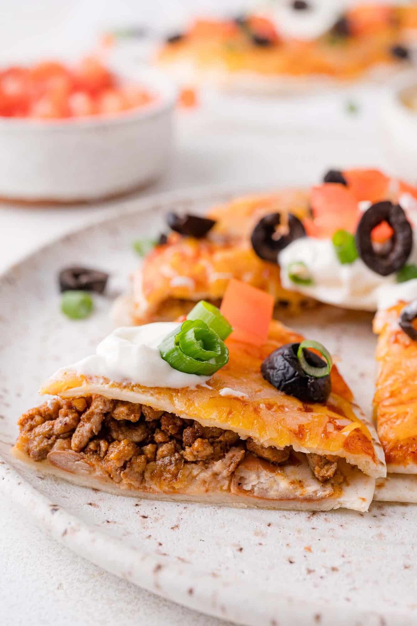 Close-up of a slice of a beef Mexican pizza topped with sour cream, diced tomatoes, sliced black olives, and green onions on a plate.