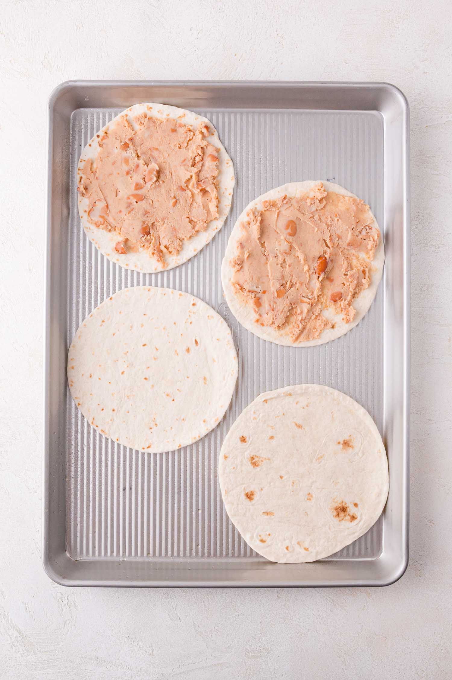 Four flour tortillas on a metal baking sheet. Two have mashed refried beans spread on top, while the other two are plain.