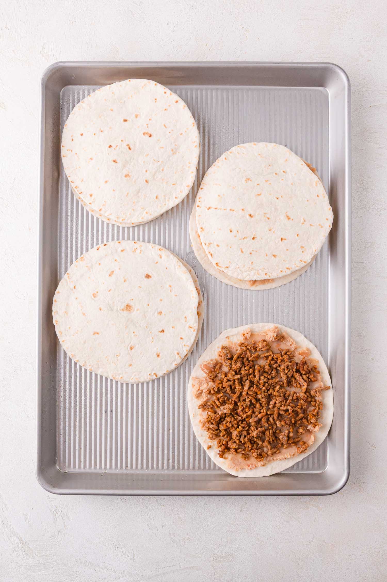Four tortilla stacks on a baking sheet, three topped with a plain tortilla and one showing cooked ground meat.
