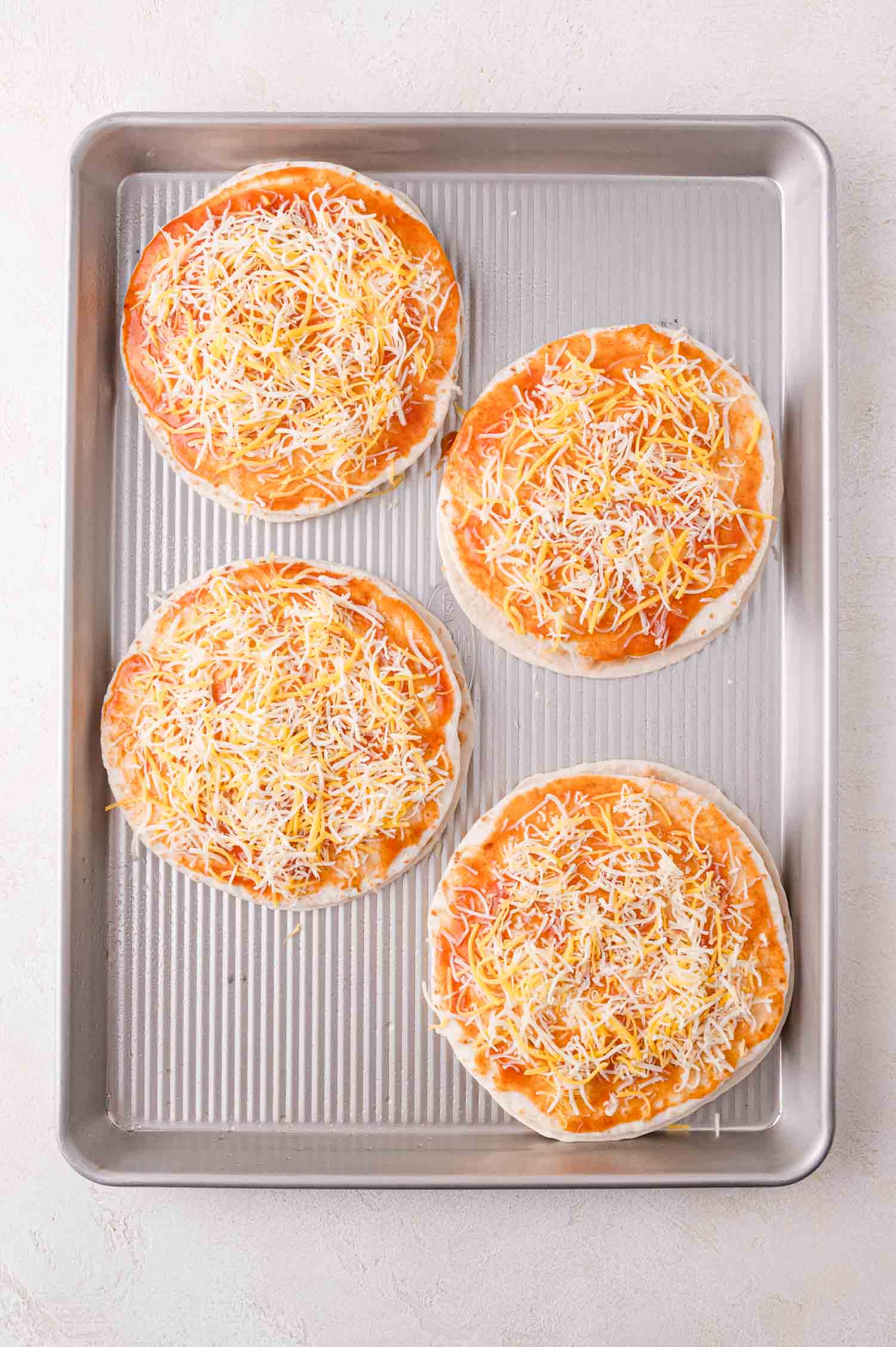 Four uncooked individual pizzas with tomato sauce and shredded cheese on a baking tray.