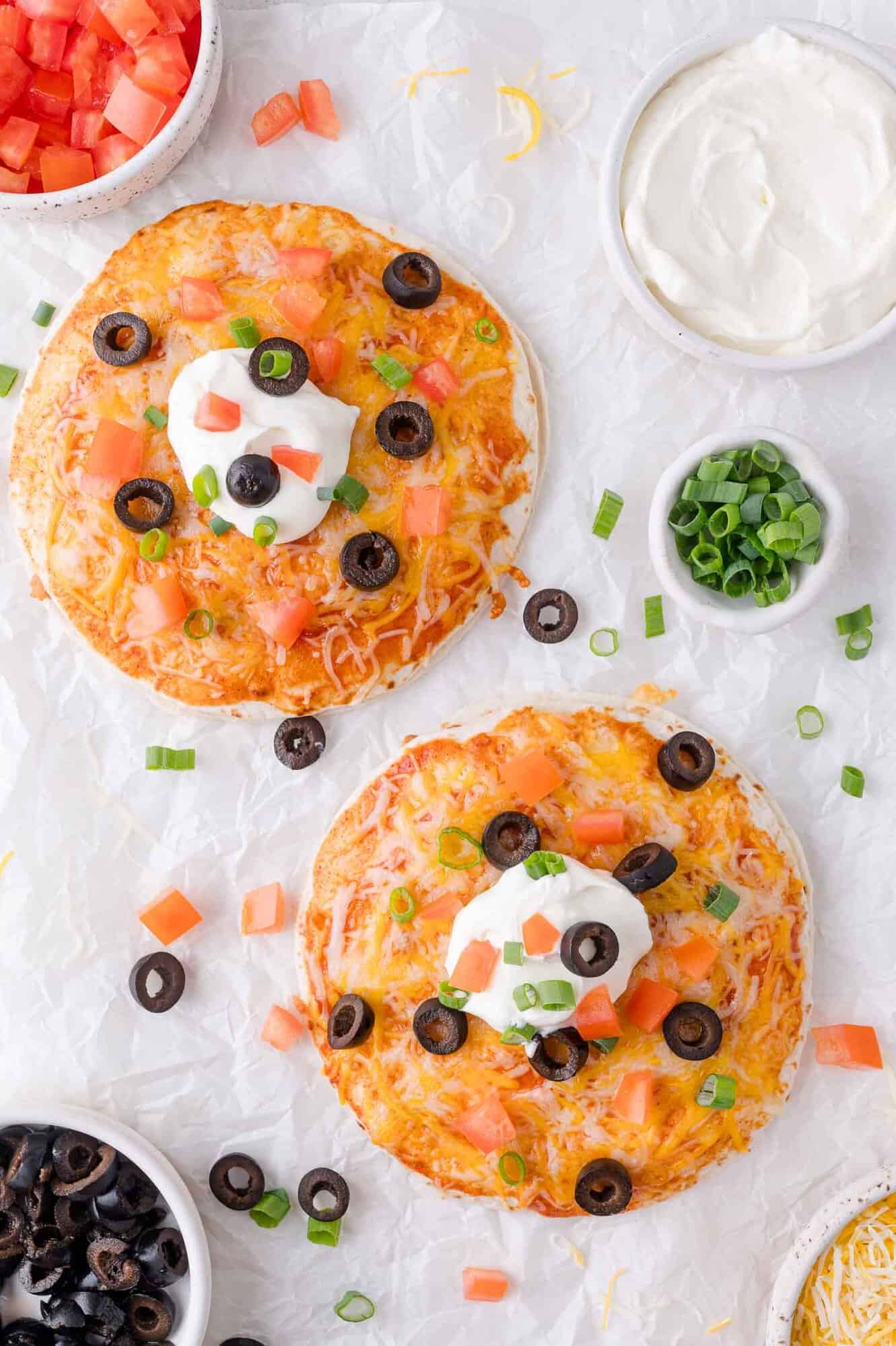 Two baked tortilla pizzas topped with melted cheese, chopped tomatoes, black olives, sour cream, and green onions on a white textured surface with small bowls of toppings nearby.
