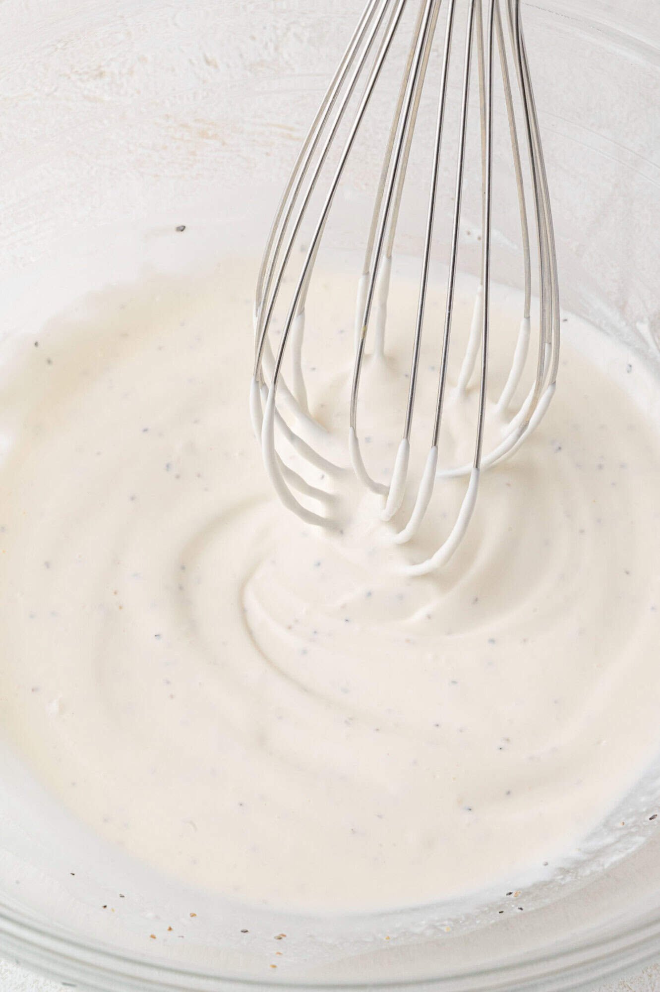 A metal whisk in a glass bowl of creamy white dressing with visible black pepper specks.