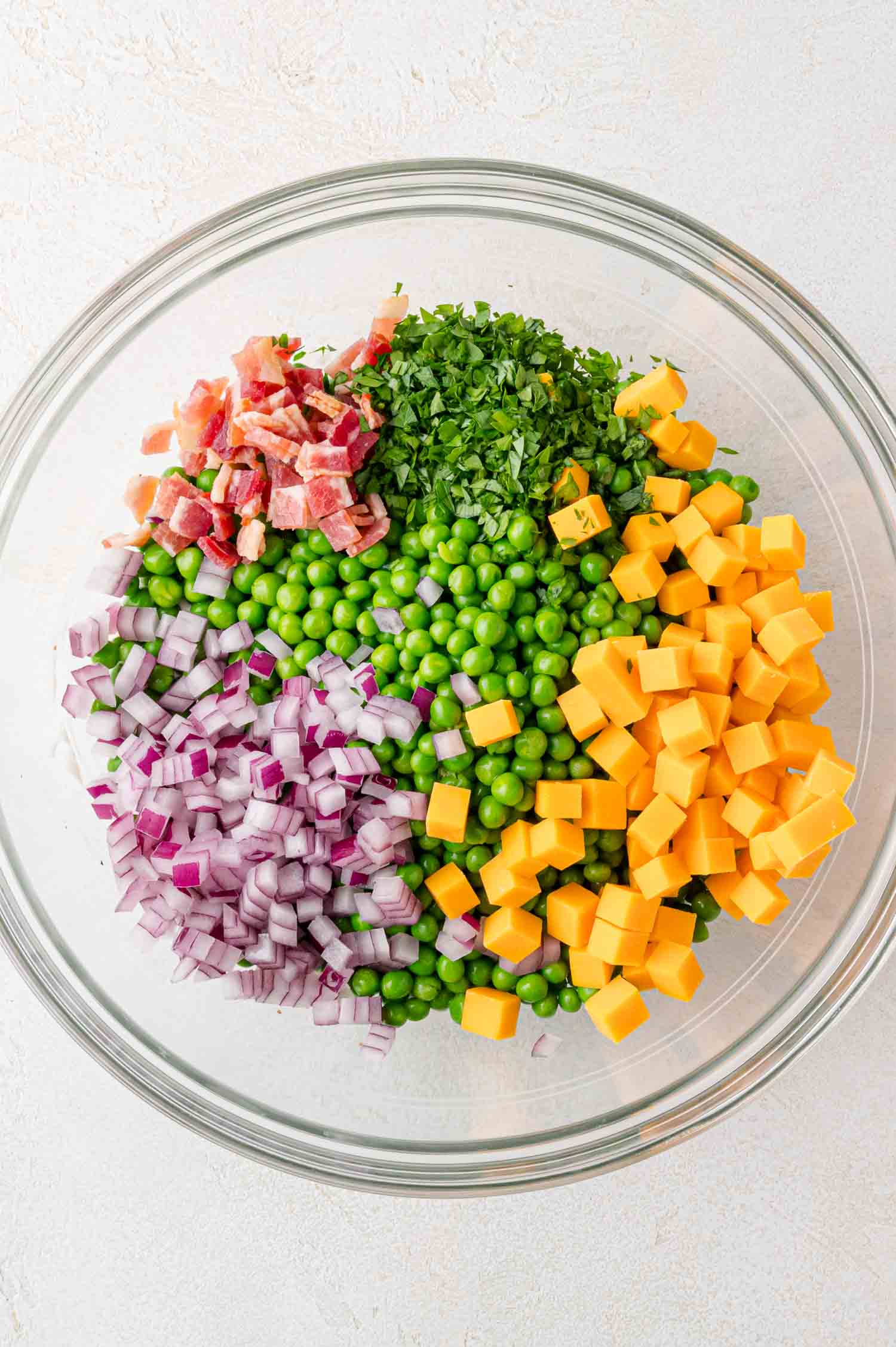 A glass bowl containing diced red onion, cheddar cheese cubes, green peas, chopped parsley, and bacon pieces, arranged in separate sections.