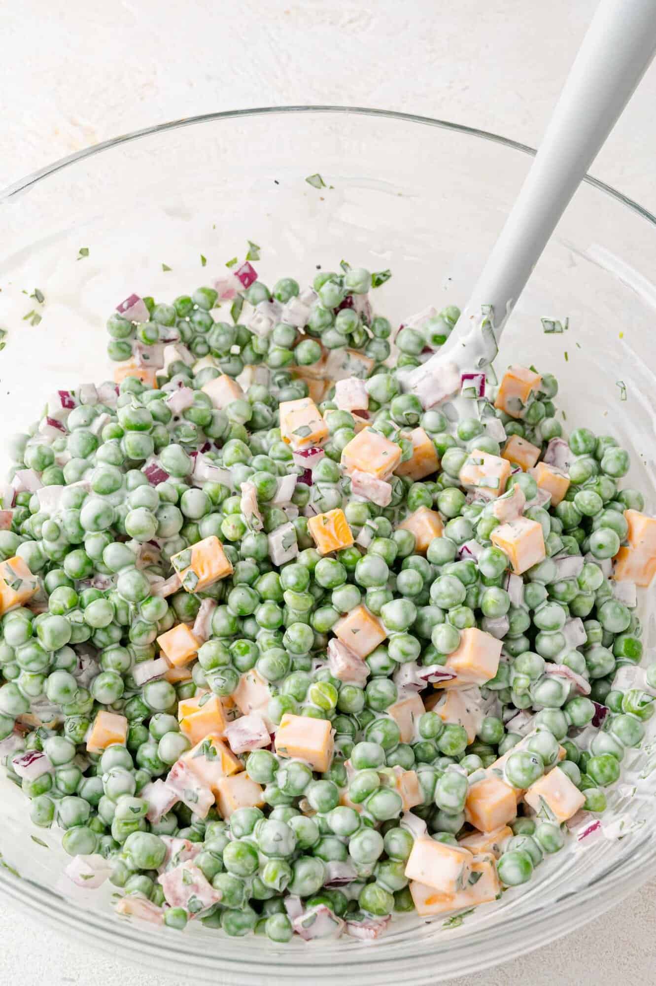 A glass bowl filled with a creamy pea salad containing green peas, cubed cheddar cheese, chopped red onion, and herbs, with a spoon for mixing.