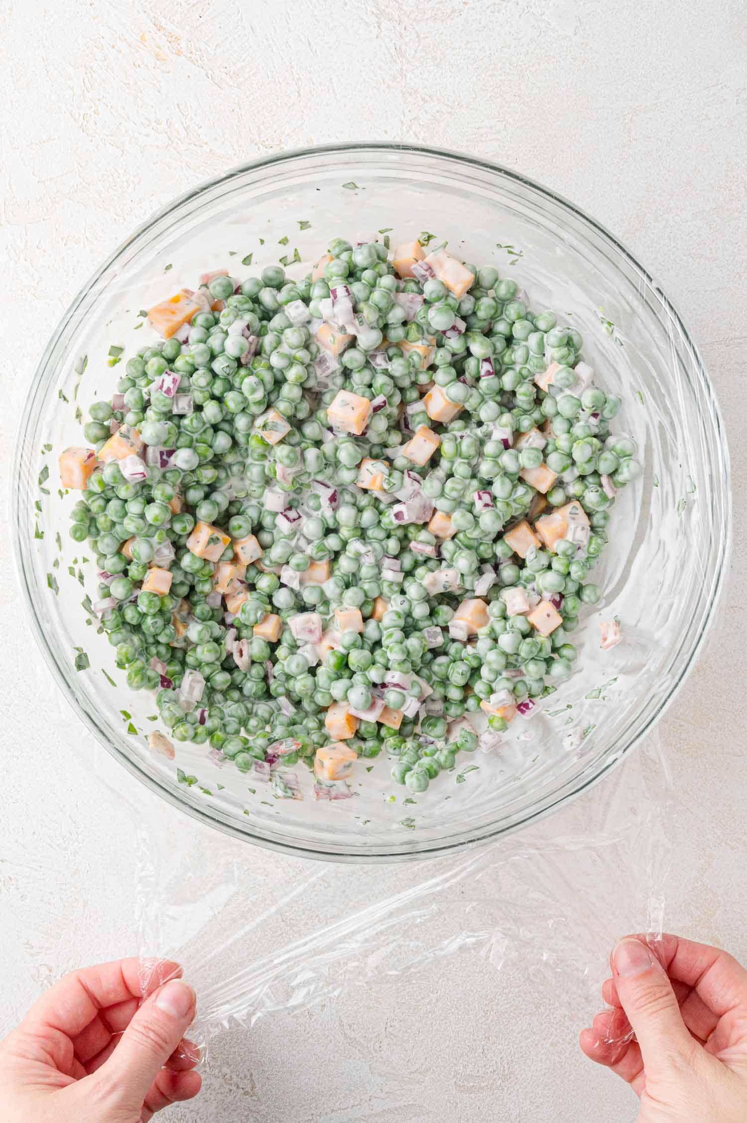 A glass bowl of pea salad with diced cheese and dressing is being covered with a piece of plastic wrap.