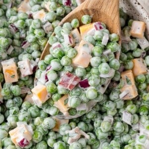 Close-up of a creamy salad with green peas, cubed cheddar cheese, chopped red onions, and bacon bits, served with a wooden spoon.