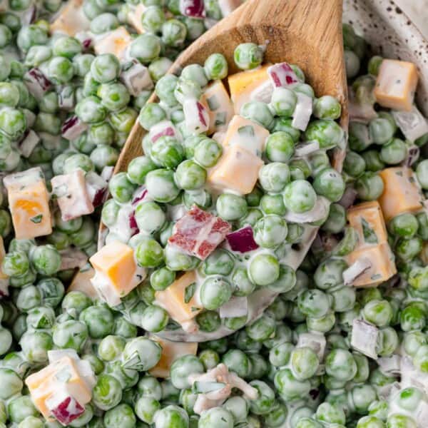 Close-up of a creamy salad with green peas, cubed cheddar cheese, chopped red onions, and bacon bits, served with a wooden spoon.