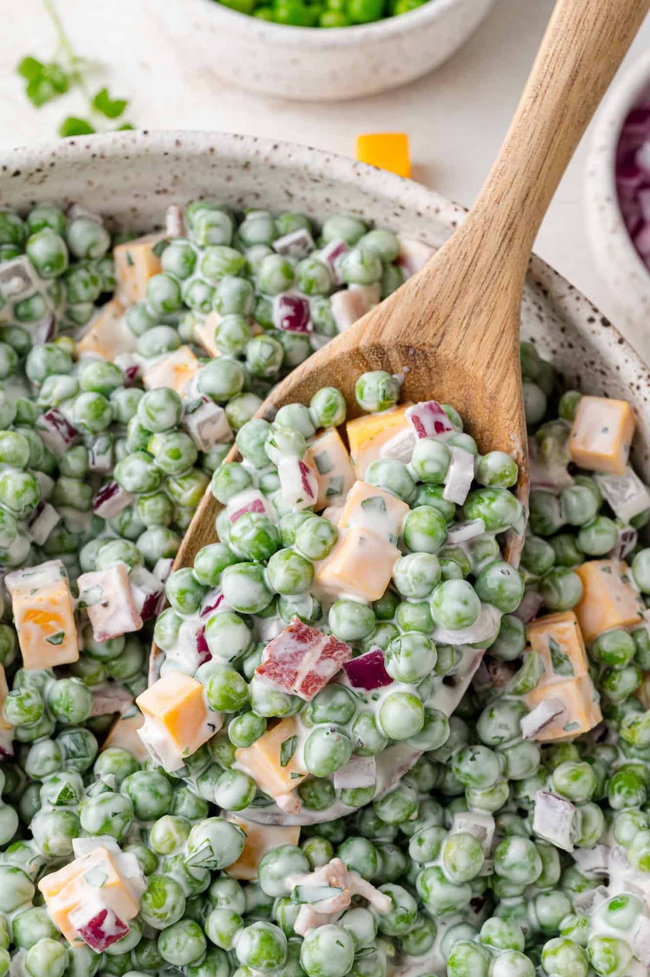 A wooden spoon scoops a creamy salad made of green peas, diced cheddar cheese, red onions, and dressing from a speckled bowl.