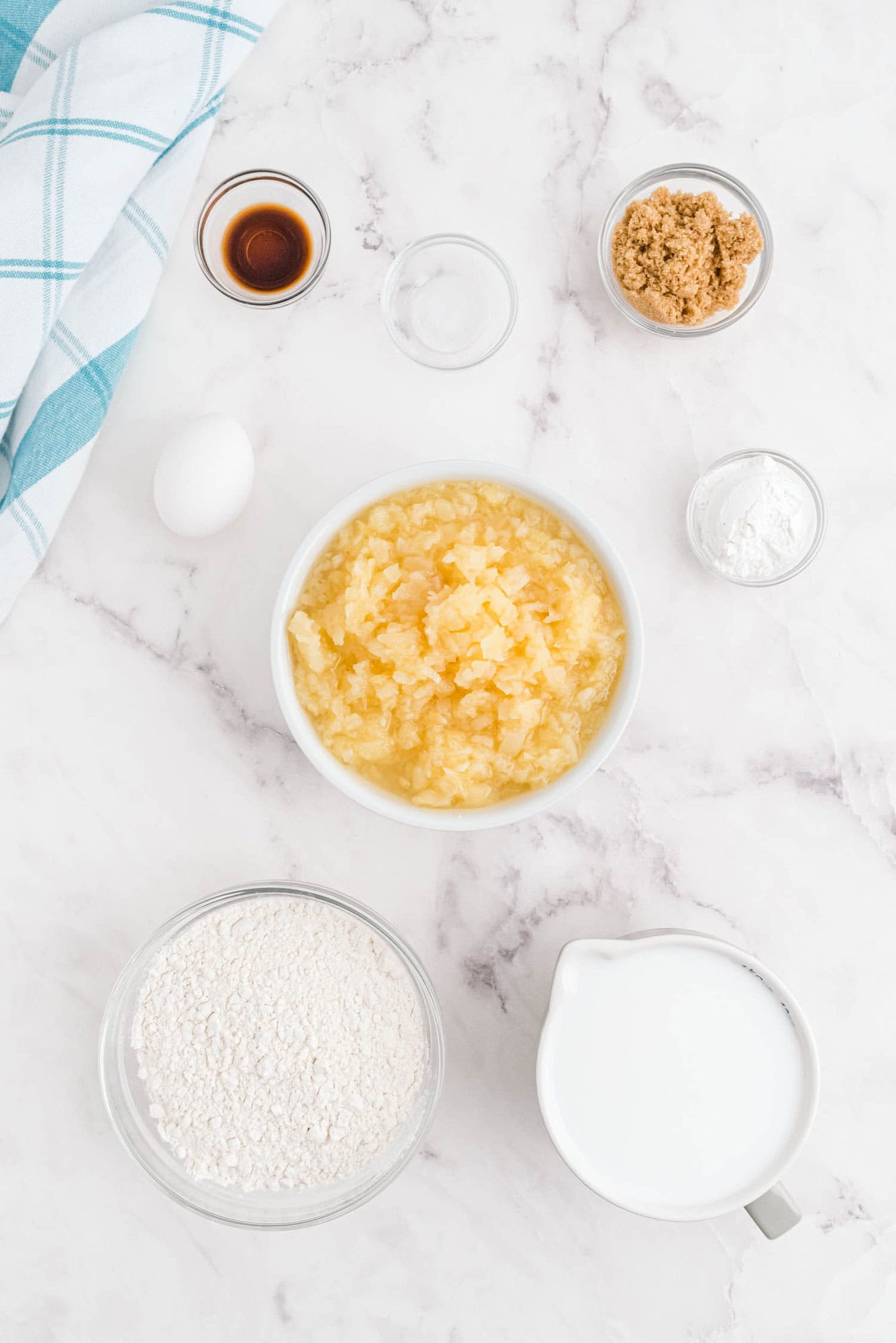 Ingredients for a recipe arranged on a marble surface: flour, milk, brown sugar, egg, vanilla, crushed pineapple, baking powder, and salt. Blue and white towel at the side.