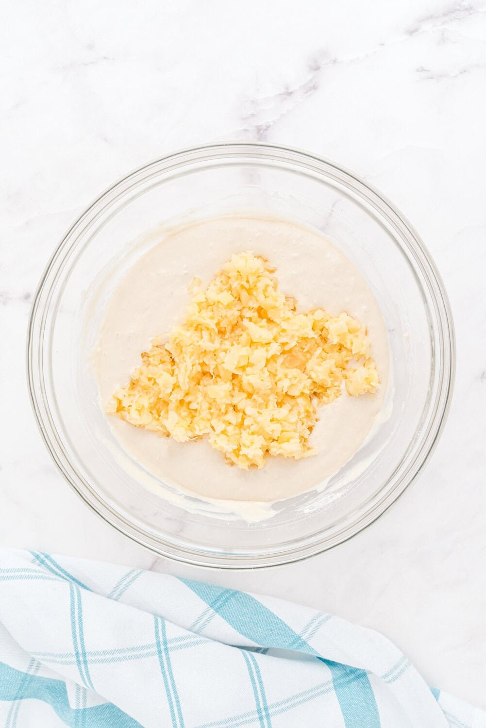 A glass bowl containing batter with chopped pineapple on top, placed on a white surface next to a blue and white checkered cloth.