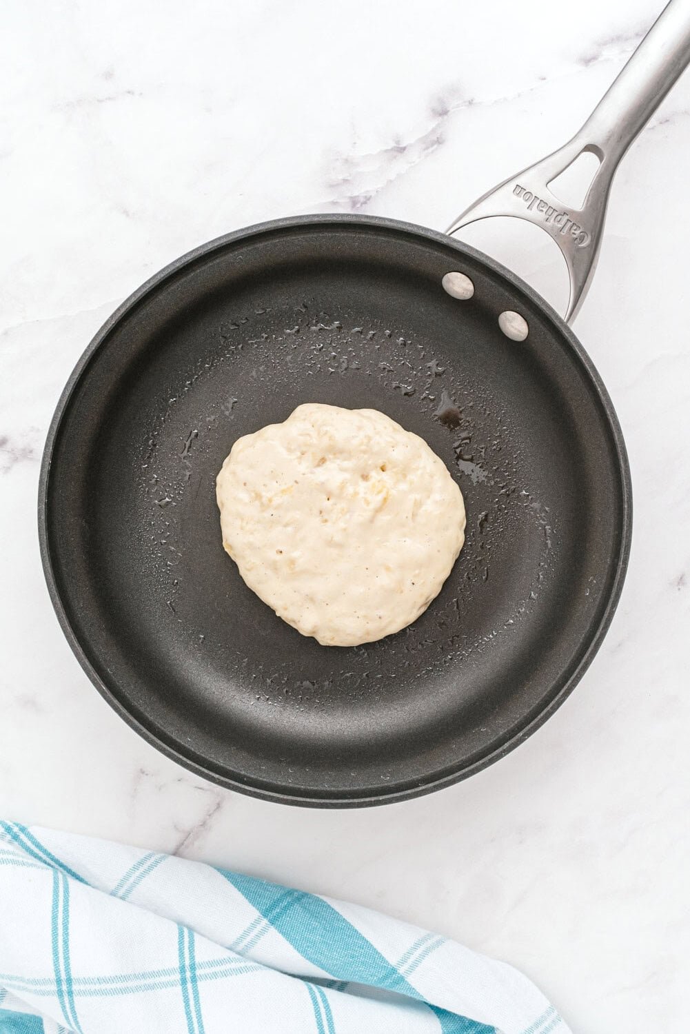 A raw pancake batter portion cooking in a nonstick frying pan on a white marble surface with a blue and white cloth nearby.
