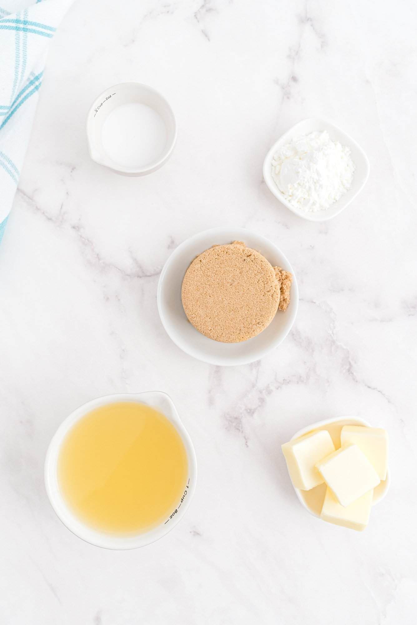 Five small bowls on a white marble surface contain milk, cornstarch, brown sugar, butter, and a cup of light brown liquid, likely broth or juice. A blue and white towel is in the corner.
