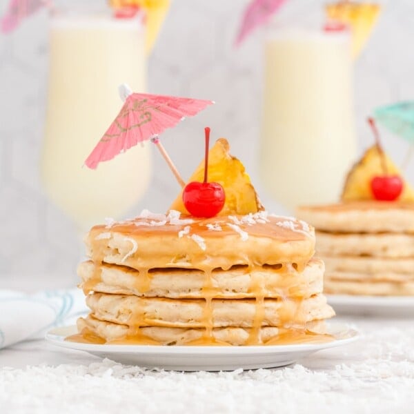A stack of pancakes topped with caramel sauce, a pineapple slice, a cherry, shredded coconut, and a paper drink umbrella, with drinks in the background.