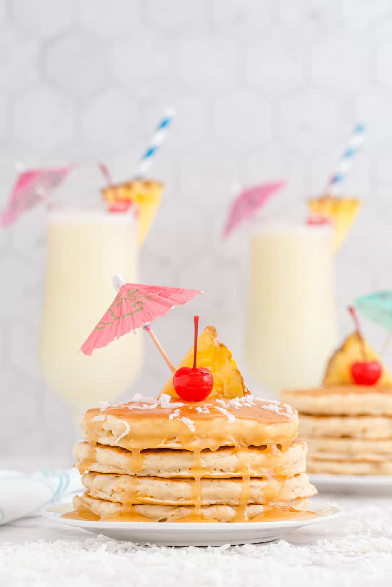 A stack of pancakes topped with syrup, shredded coconut, a pineapple slice, a cherry, and a paper umbrella, with two piña colada drinks in the background.