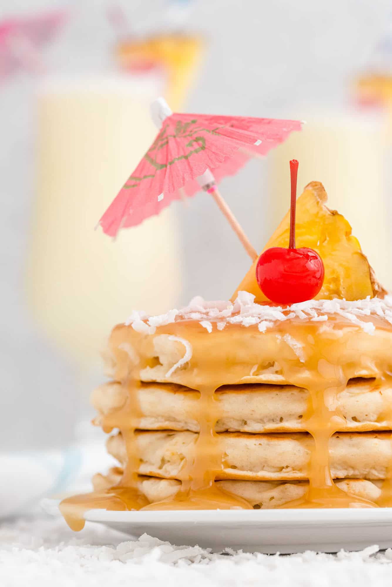 A stack of pancakes topped with caramel sauce, shredded coconut, a pineapple slice, a maraschino cherry, and a pink paper umbrella.