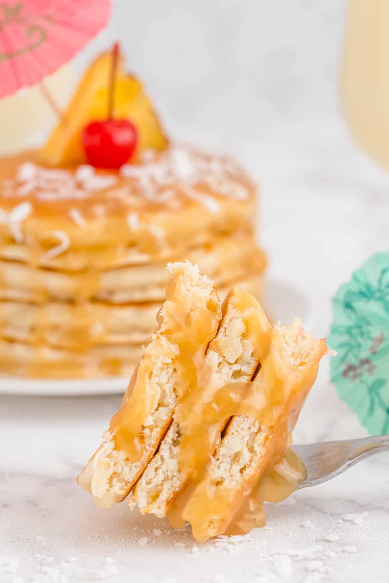 A fork holds a bite of pancakes drizzled with syrup, with a stack of decorated pancakes and cocktail umbrellas in the background.