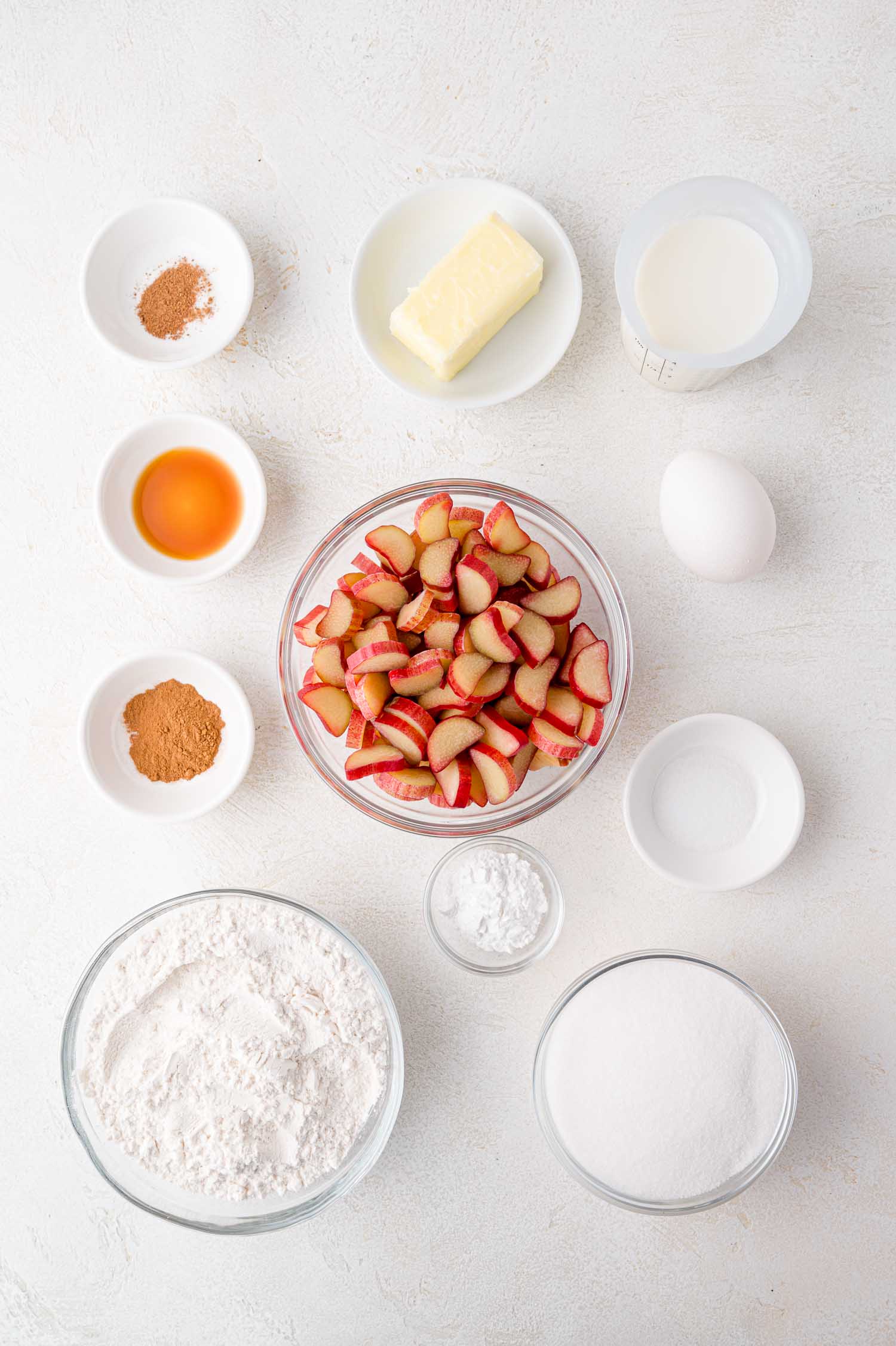Bowls and plates of baking ingredients including chopped rhubarb, flour, sugar, butter, milk, an egg, vanilla, cinnamon, nutmeg, baking powder, and salt arranged on a white surface.