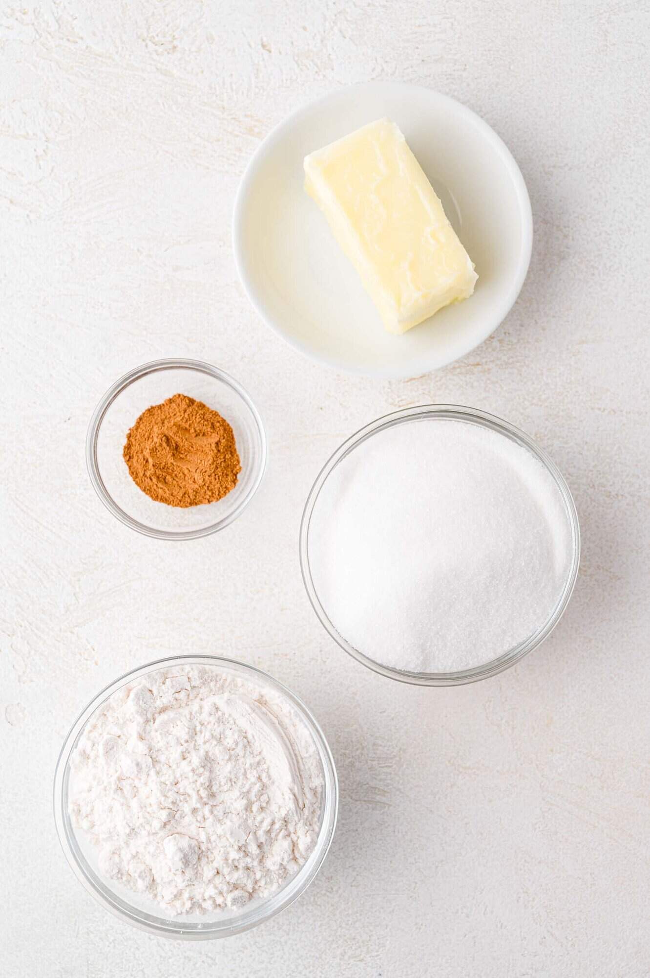 A stick of butter on a plate, a small bowl of cinnamon, a bowl of sugar, and a bowl of flour arranged on a white surface.