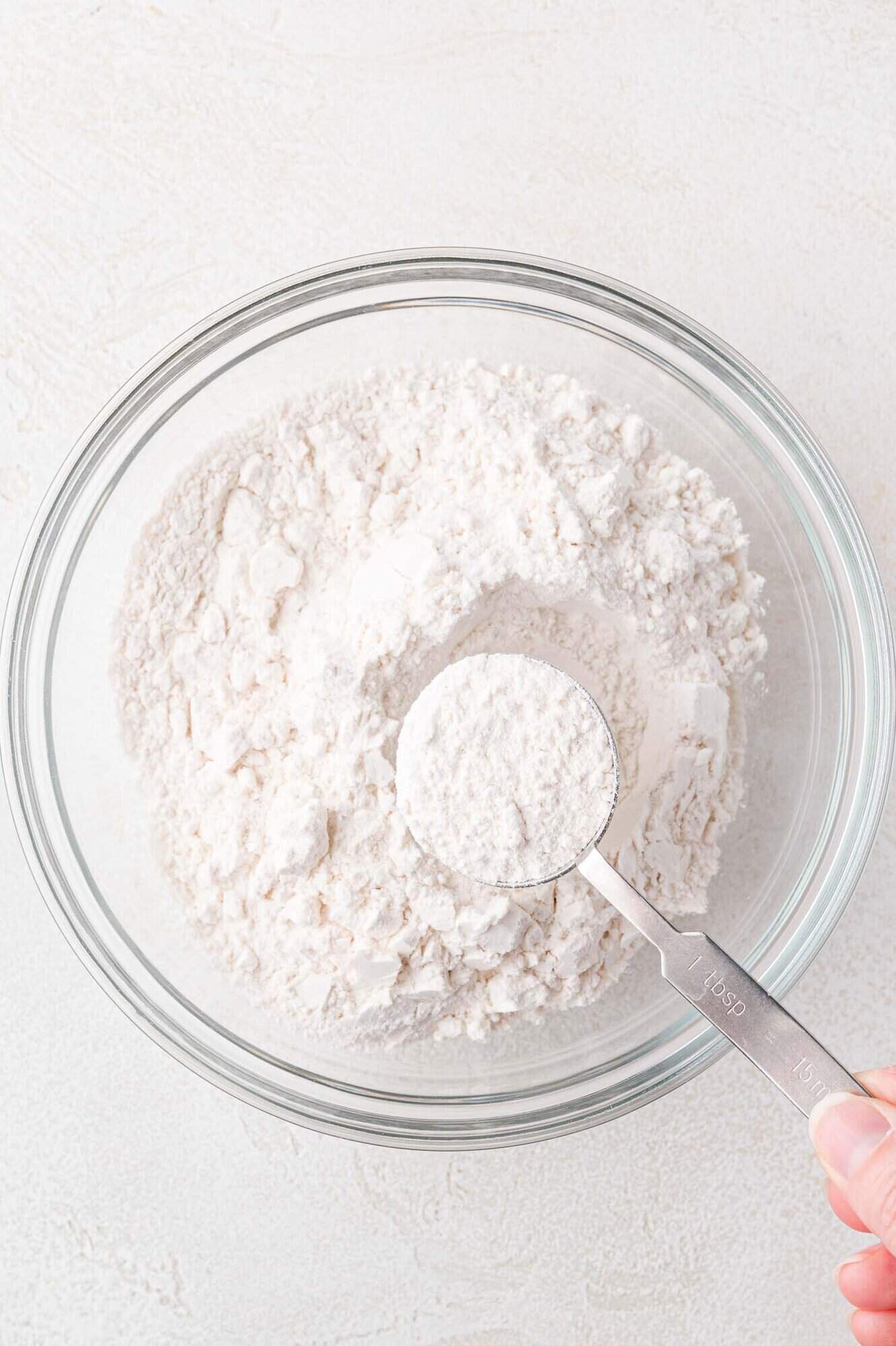 A glass bowl filled with flour with a metal measuring spoon scooping some flour from the bowl on a light surface.