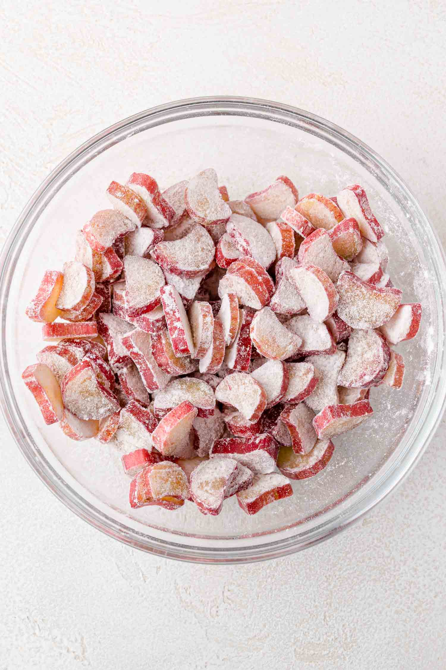 A glass bowl filled with sliced rhubarb coated in granulated sugar and flour, placed on a light-colored surface.