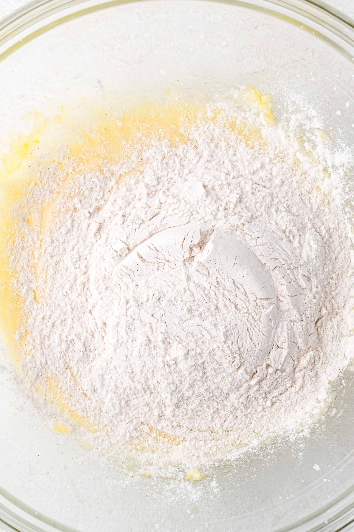 A glass mixing bowl containing flour on top of a yellow, partially mixed batter.