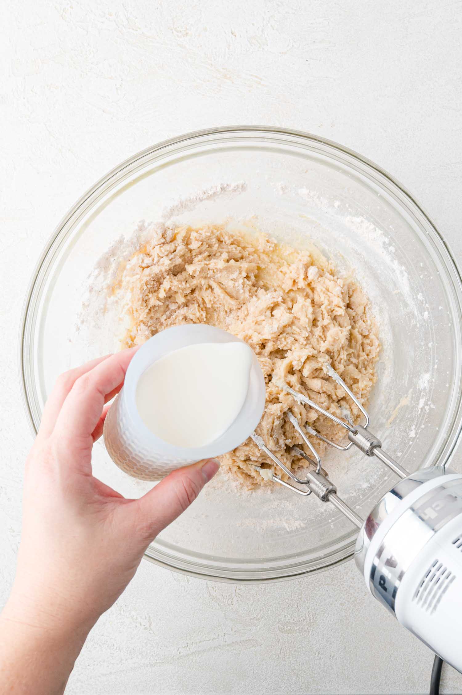 A hand holds a cup of milk above a mixing bowl containing dough, with an electric mixer resting in the bowl.