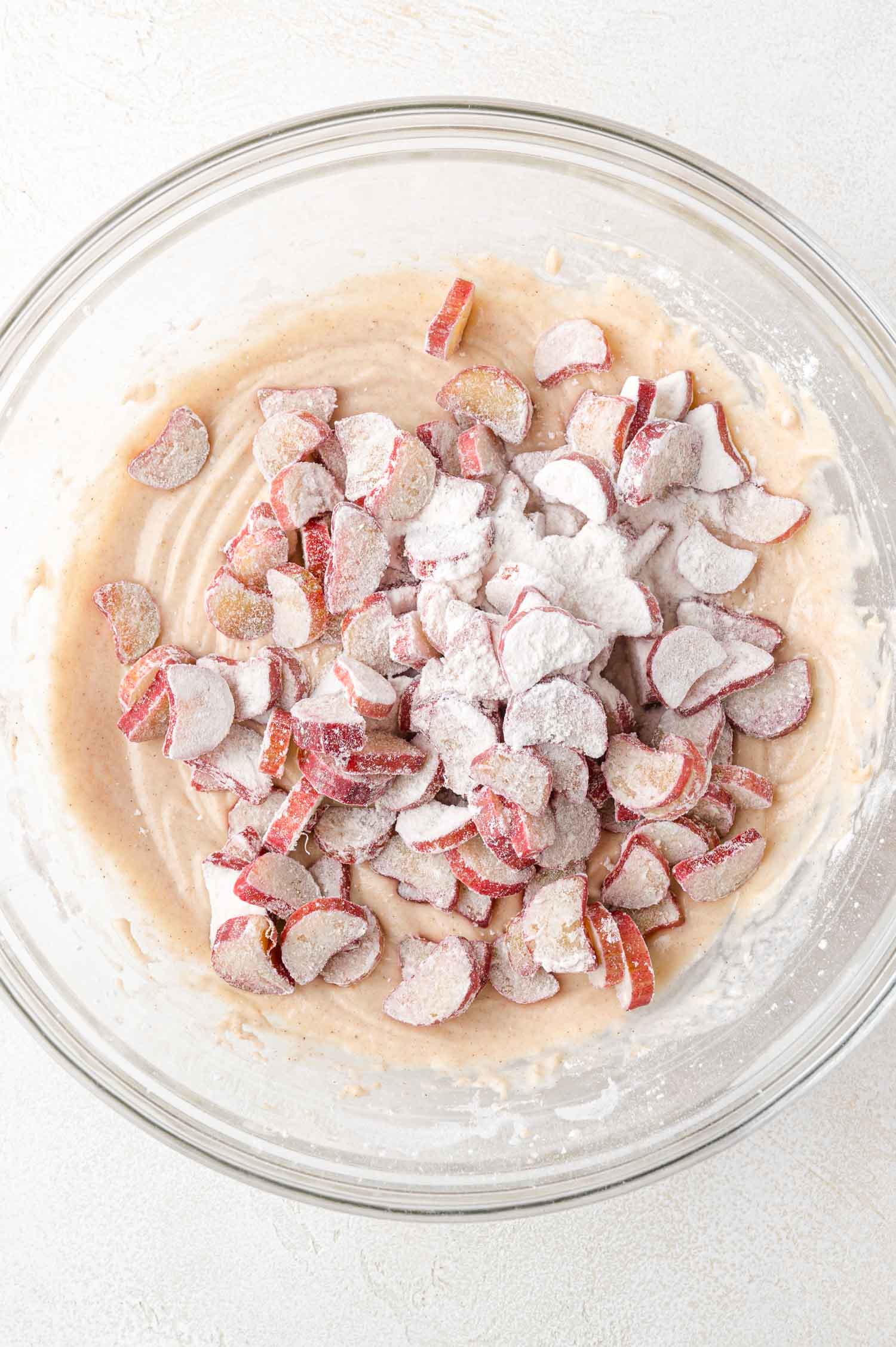A glass bowl contains cake batter mixed with sliced rhubarb pieces coated in flour, sitting on a light-colored surface.