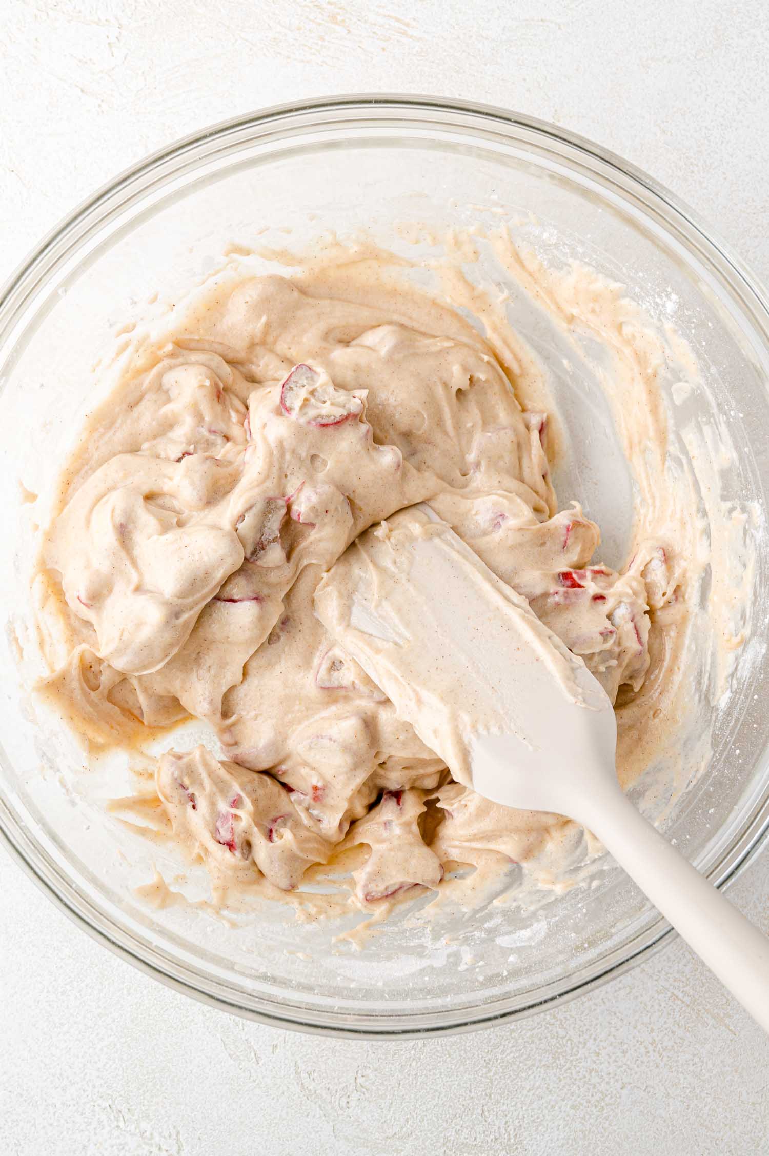 A glass bowl filled with thick, beige batter containing red fruit pieces, being mixed with a white spatula on a light surface.