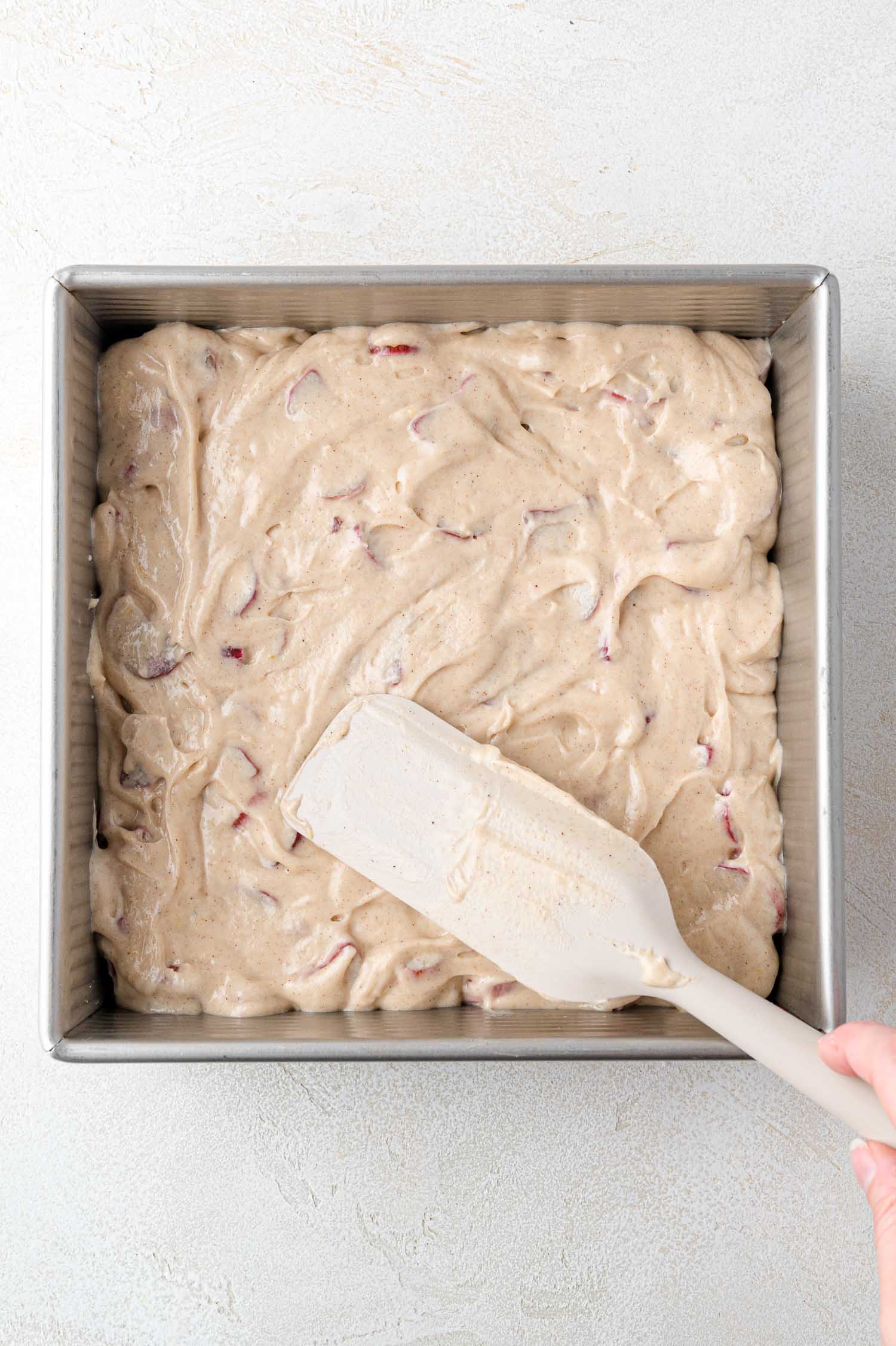 A hand holding a spatula spreads cake batter with visible rhubarb pieces evenly in a square metal baking pan.