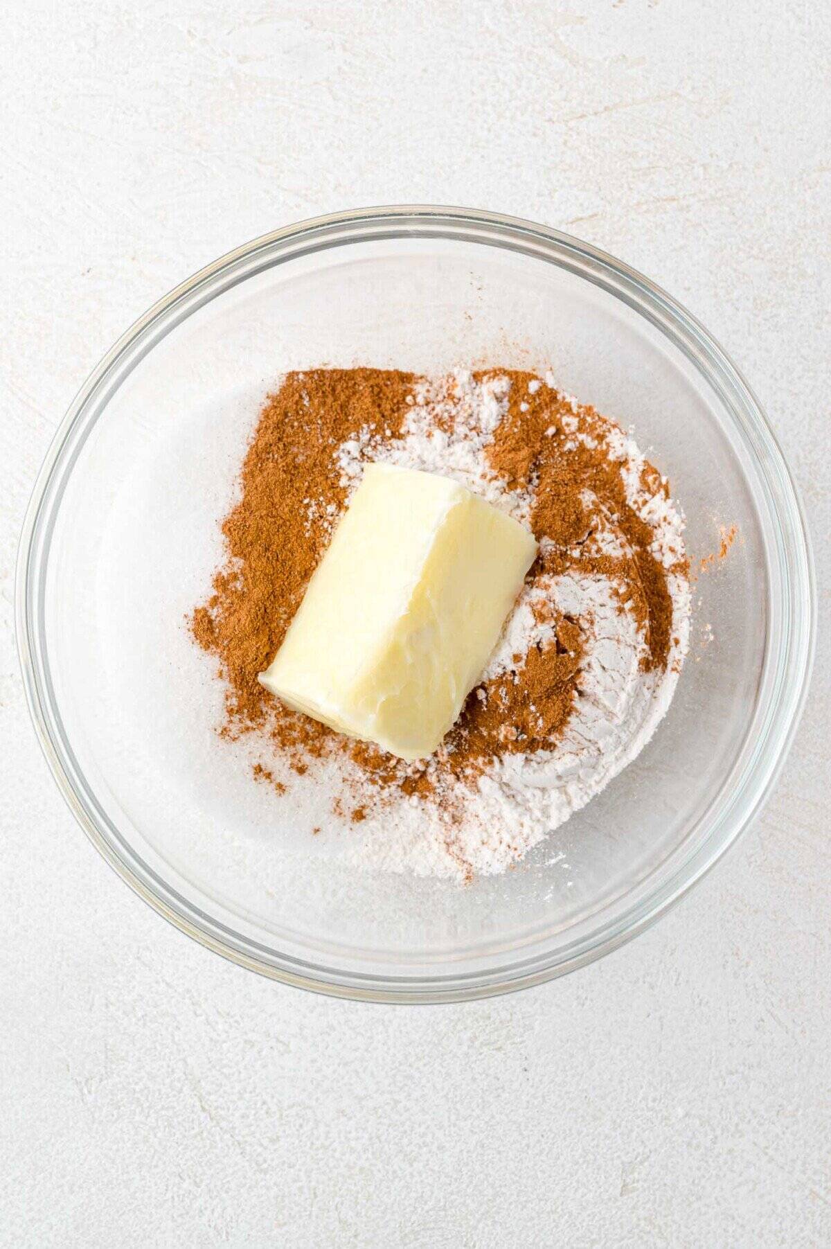 A glass bowl containing a stick of butter, cinnamon, flour, and sugar on a white surface.