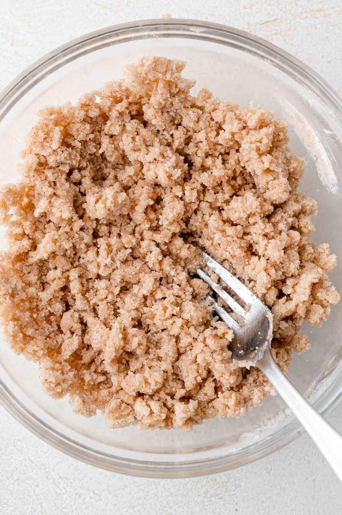 A glass bowl filled with a crumbly brown sugar and cinnamon mixture, with a metal fork resting in the mixture.