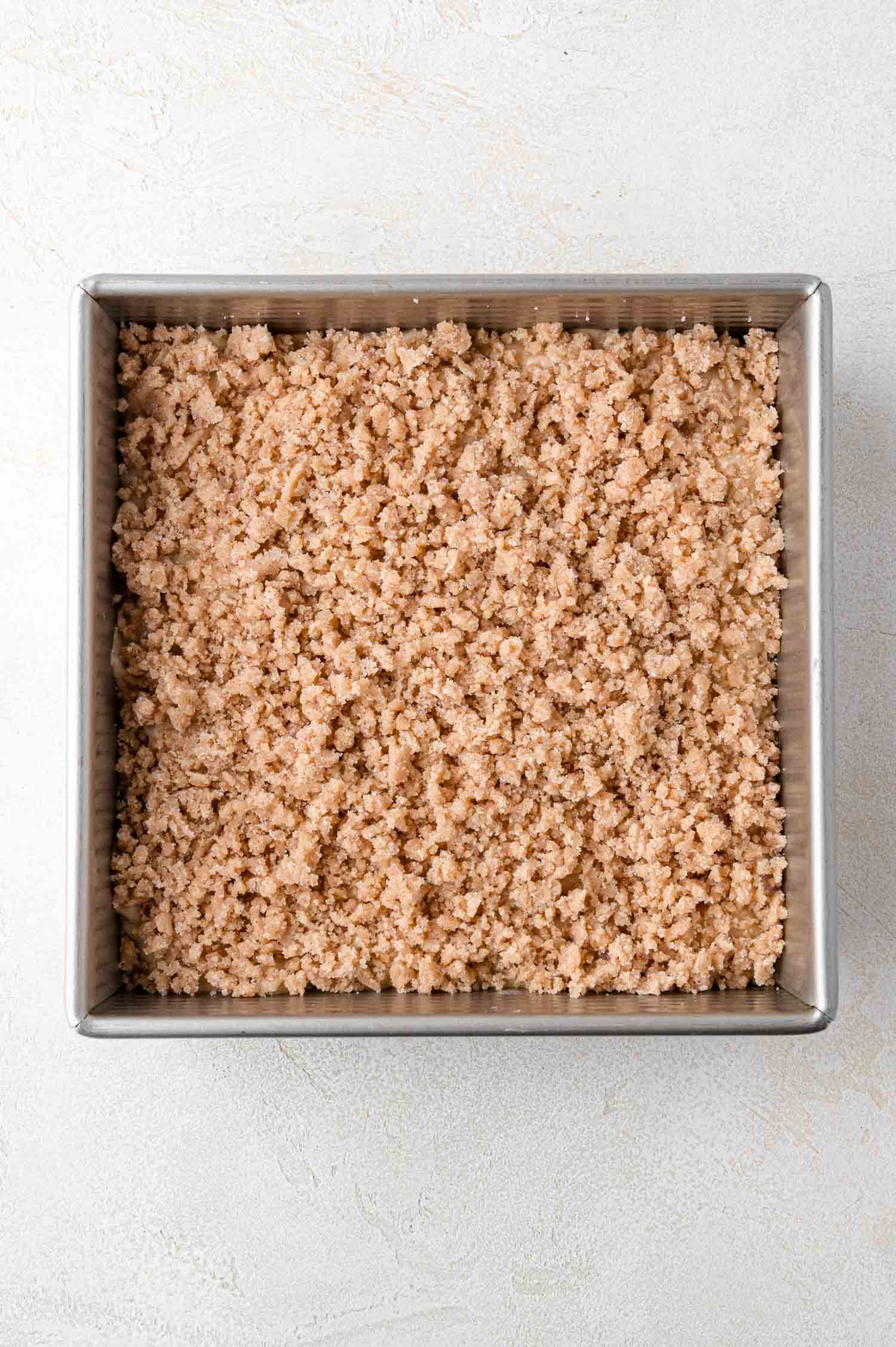 A square metal baking pan filled with an even layer of unbaked crumb topping on top of a cake on a light surface.