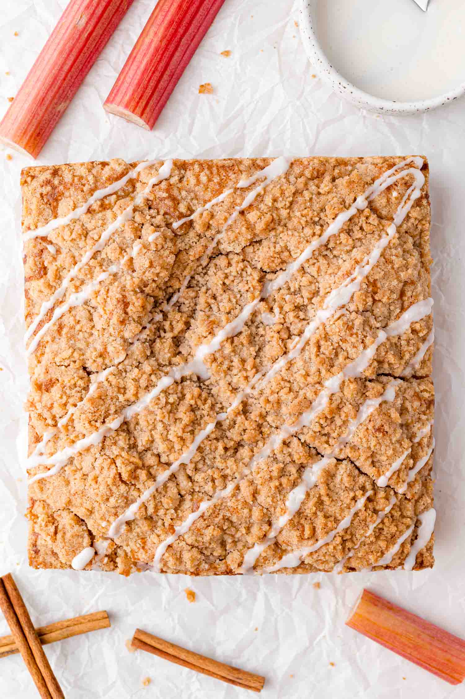 Square crumb cake with white icing drizzle, placed on crumpled parchment, surrounded by rhubarb stalks, cinnamon sticks, and a bowl of glaze.