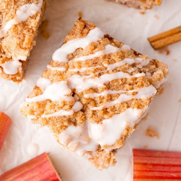A square piece of rhubarb cake with icing drizzle on top, surrounded by pieces of rhubarb and a cinnamon stick on a white surface.