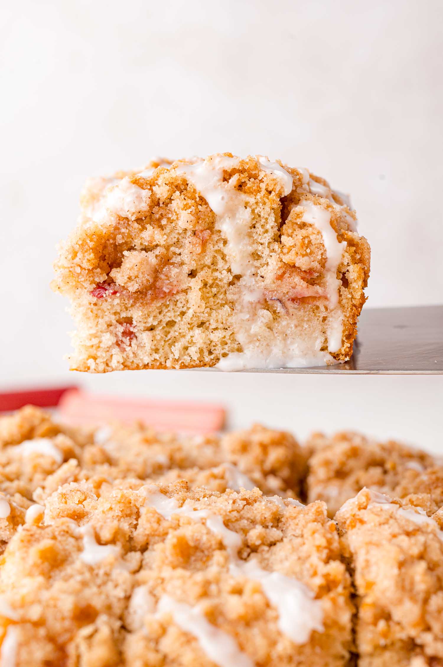 A close-up of a piece of crumb cake with a drizzle of white icing, resting above a pan of similar cake pieces.