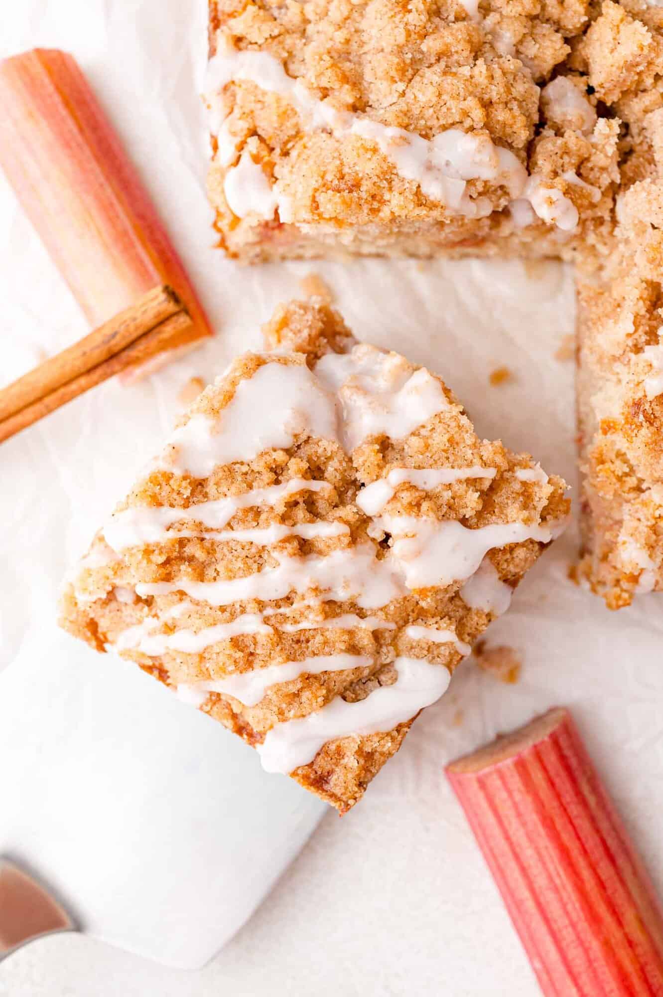 A square piece of crumb-topped coffee cake with icing drizzle, surrounded by fresh rhubarb stalks on a white surface.